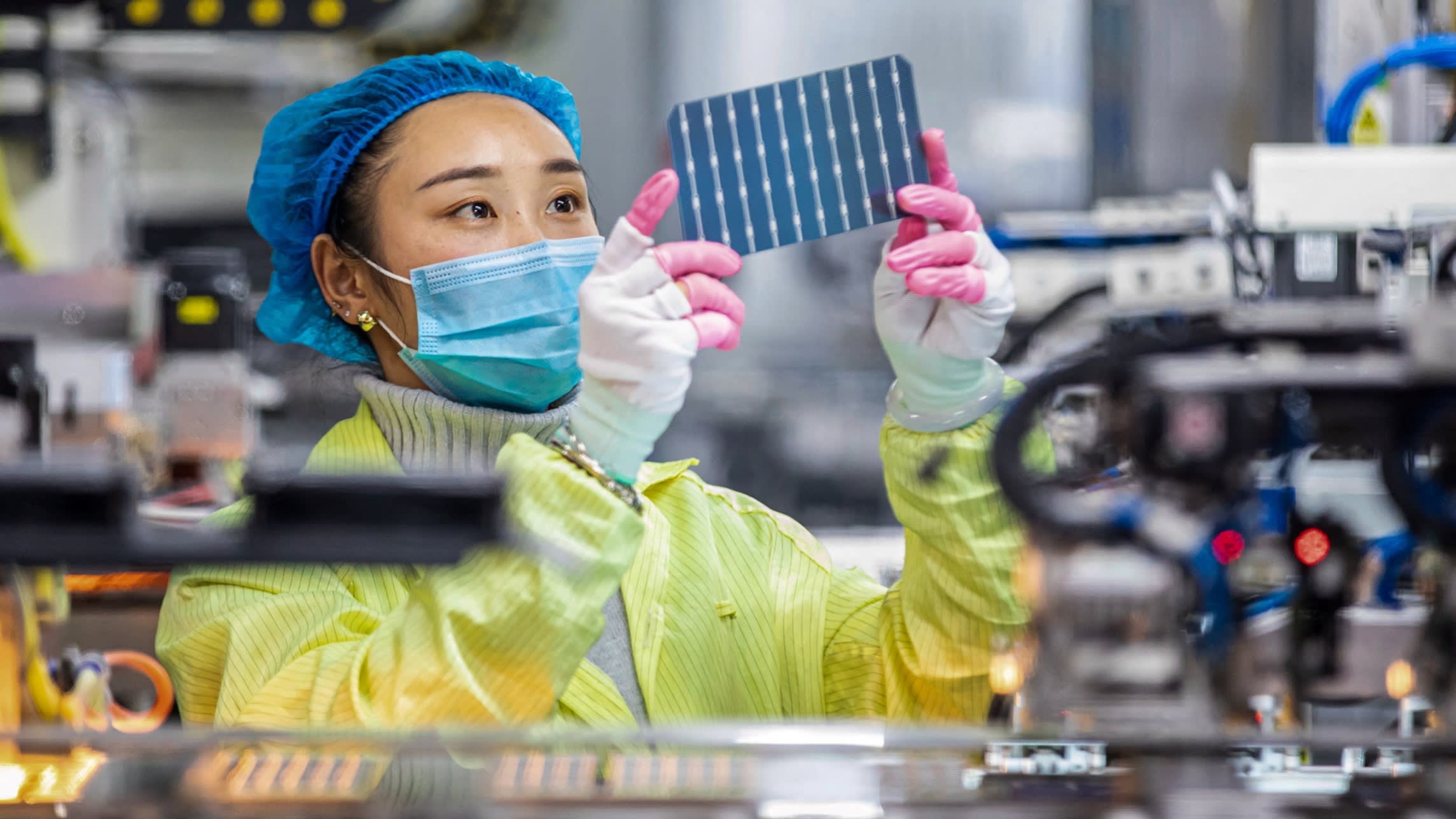 An Asian woman in hairnet, mask and plastic coverall inspects a solar photovoltaic module inside a factory