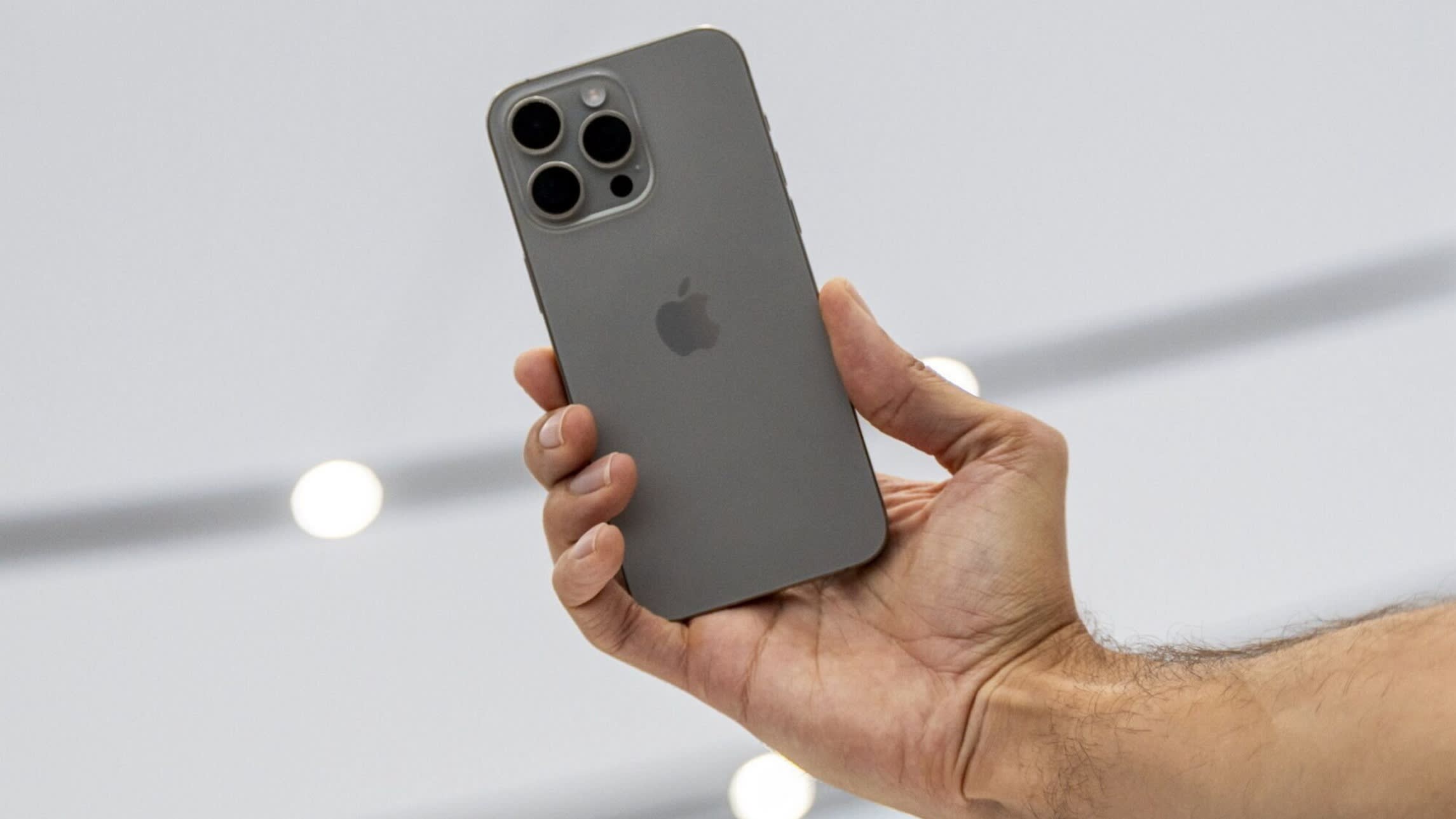 A close-up of a person’s right hand holding the iPhone 15 Pro Max showing the rear with the camera lenses and Apple logo