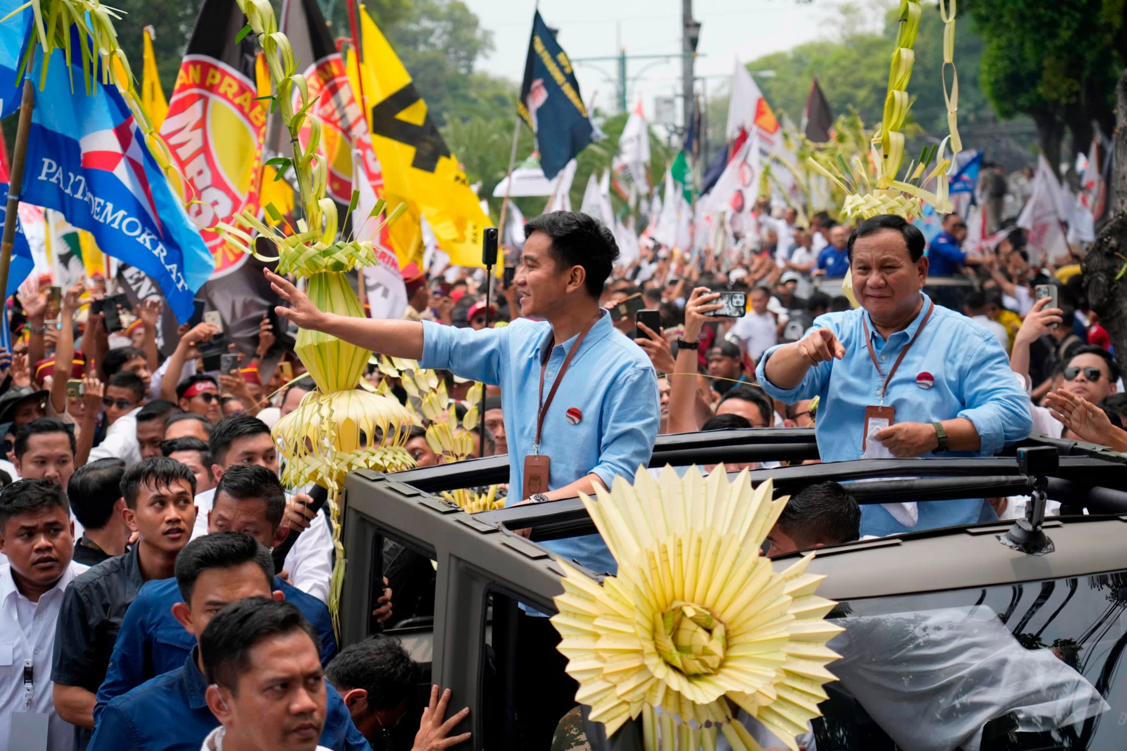 Two men in blue shirts stand waving from an open-top car as they are thronged by crowds of people
