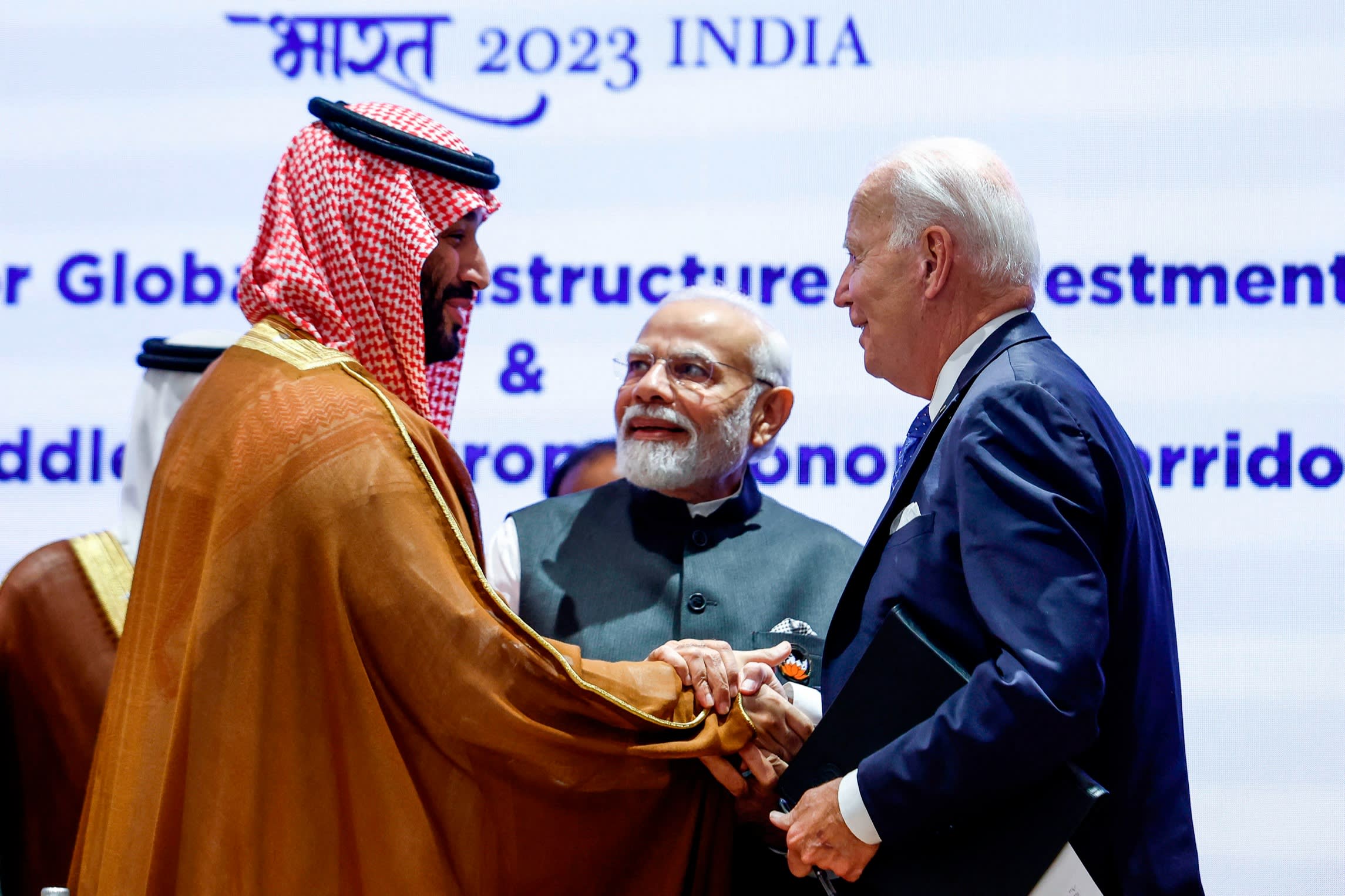 A man wearing Arabic headdress and gown shakes hands with a middle-aged man in a blue suit as a man in a green-blue suit