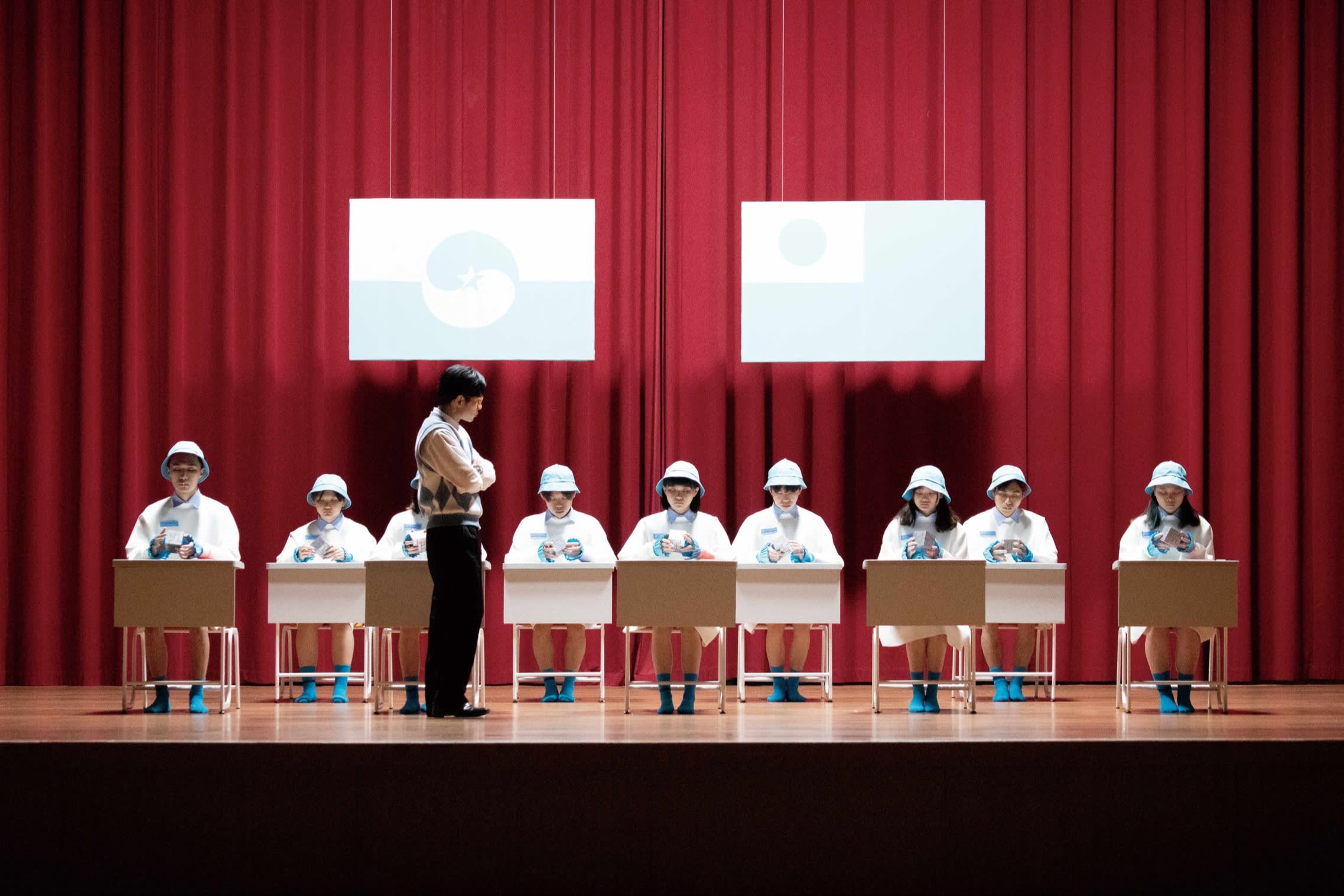 A film still of showing young students at desks arranged on a stage with a red curtain behind them. A man who looks like their teacher stands over them. Two large blue and white posters hang over their heads. 