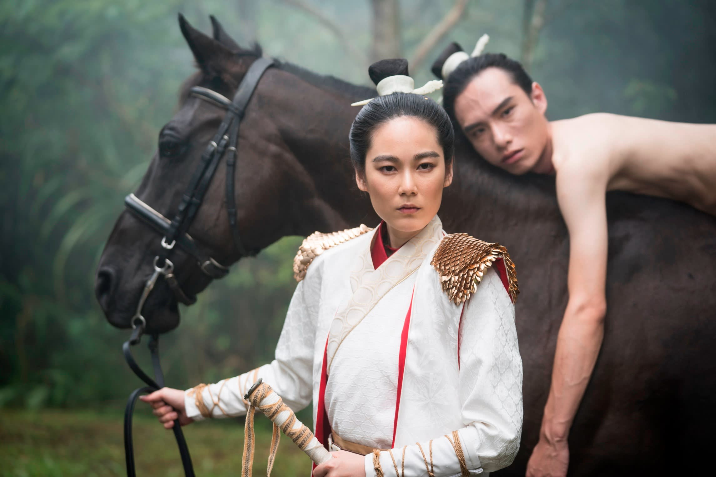 A film still of a woman in a white robe and hair in a bun holds the reins of a horse while a man with the same hairstyle lies on top of the horse.