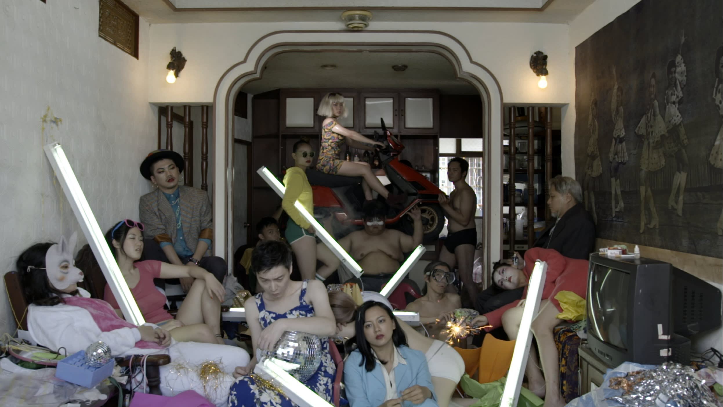 A film still showing a group of people, some dressed, some half naked, sit on the floor of a living room or pose in the doorway holding LED bars.
