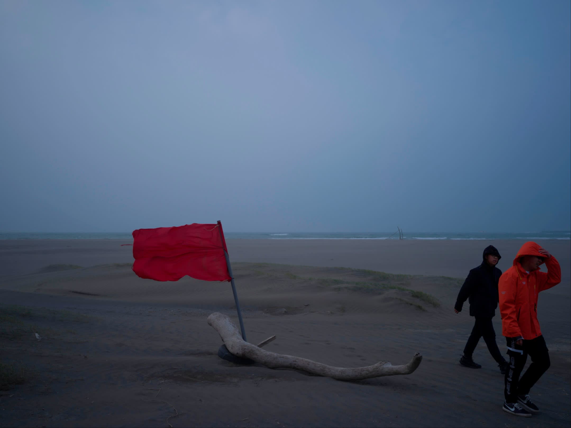 Two young men in waterproof jackets walk past a red flag on a blustery beach