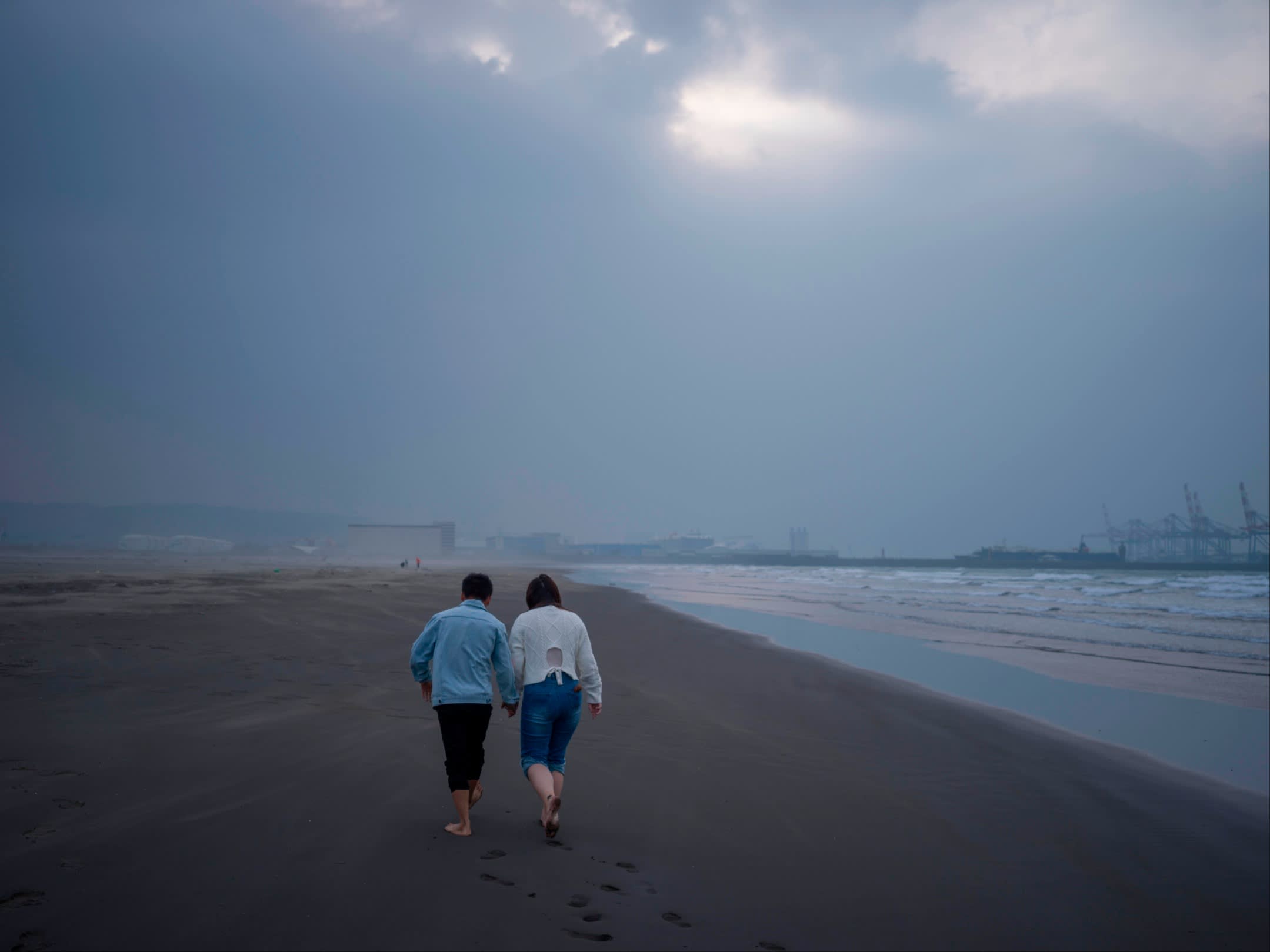 A man and a woman, seen from behind, walk hand in hand along a beach on an overcast day