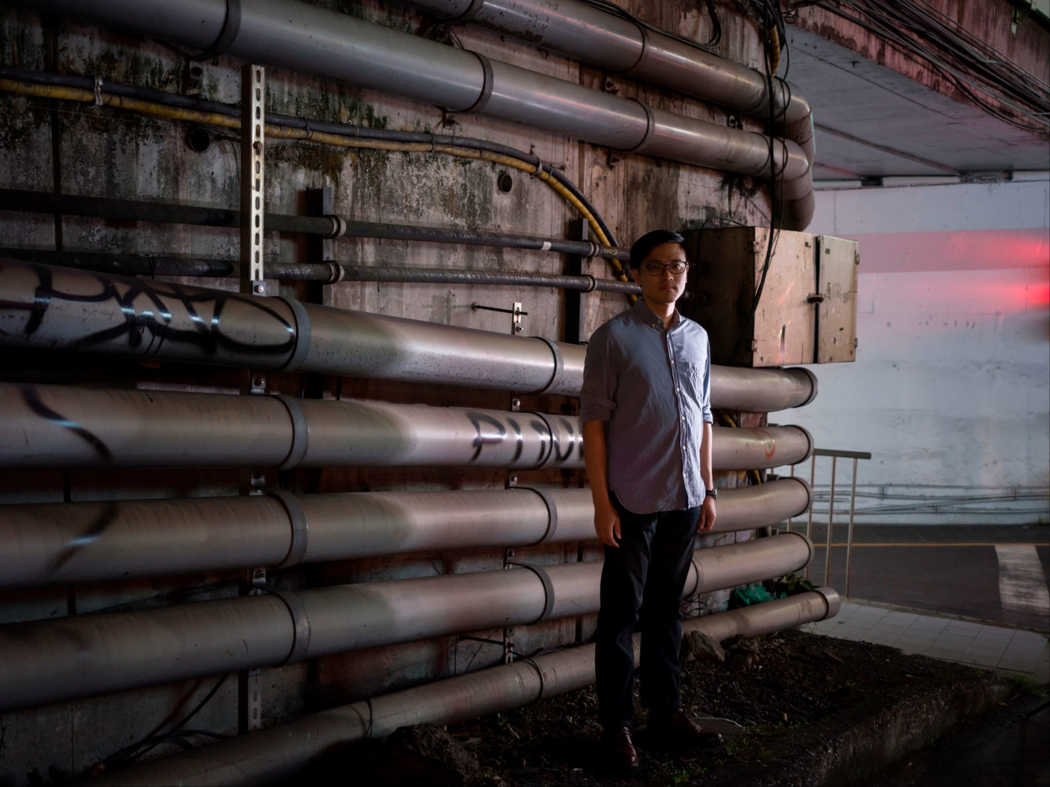 A young man in glasses and blue shirt stands in front of a wall lined with pipes