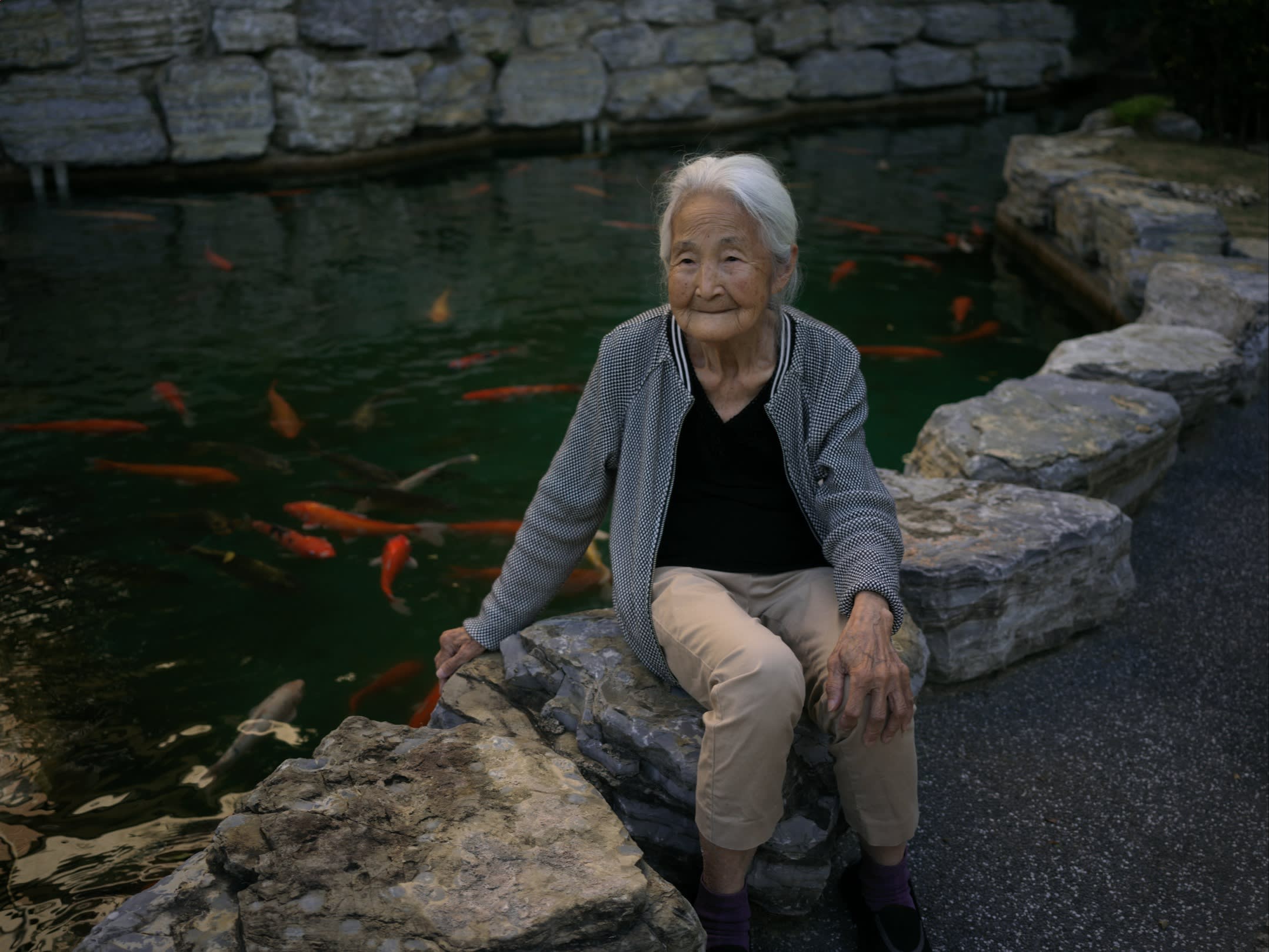 A grey-haired woman sits by the side of a carp pond
