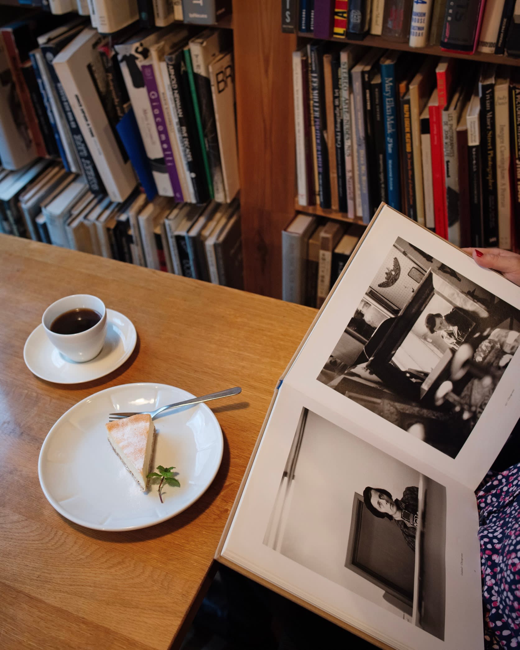 An open photography book beside a slice of tart on a plate and a cup of coffee, all on a wooden table, at Megutama