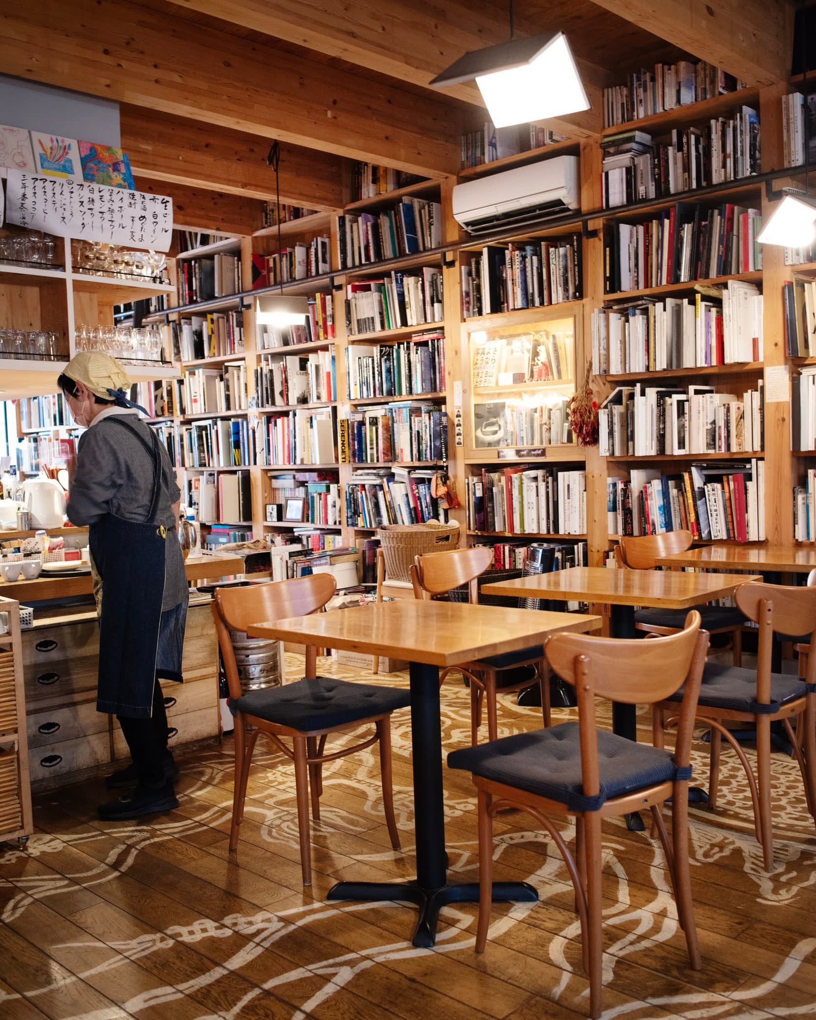 A long book-filled wall at Megutama, with a female member of staff and tables and chairs in the foreground