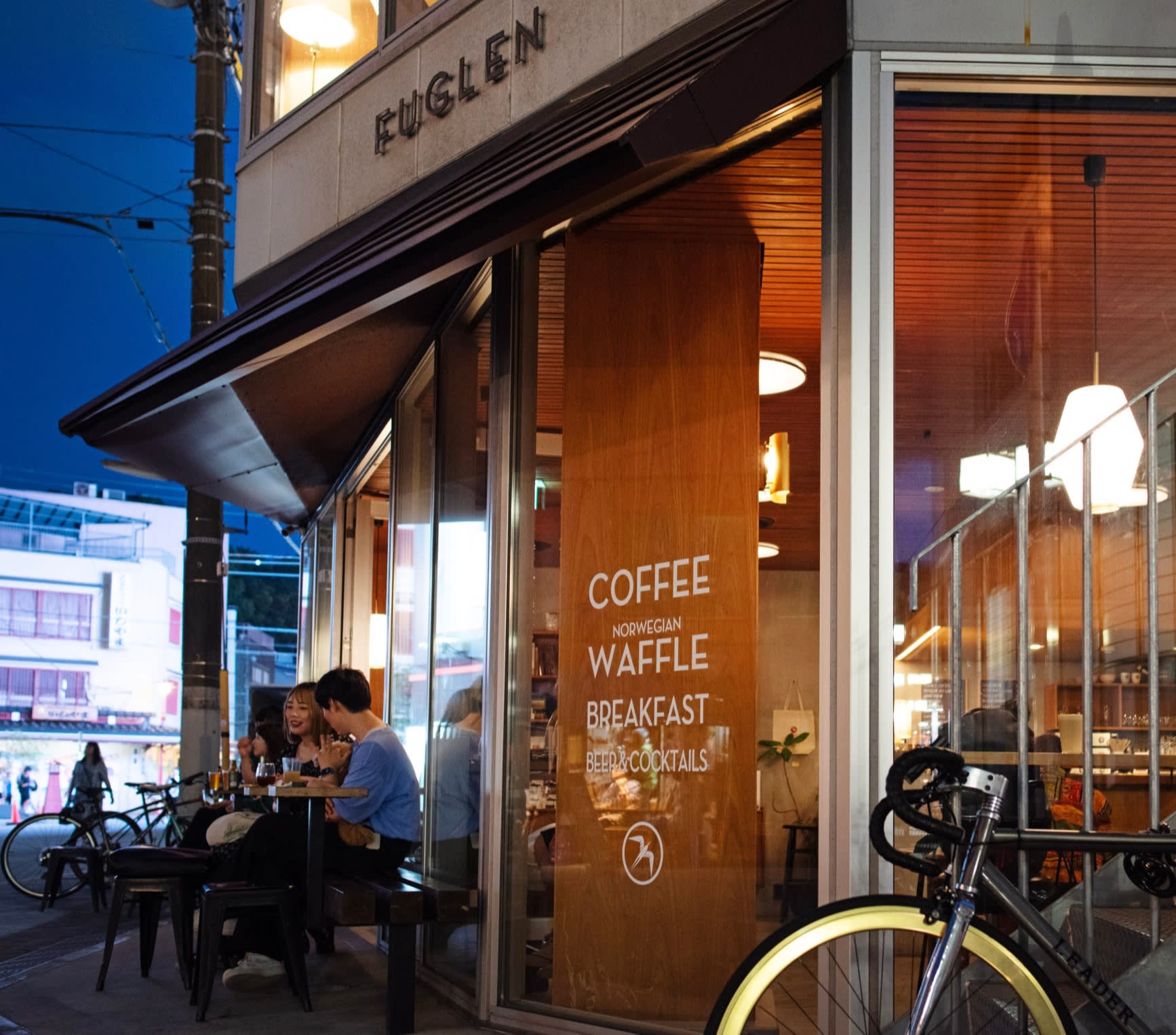 A man and a woman at table outside Fuglen Asakusa in the evening, with its Scandinavian-style wooden interior visible through the windows