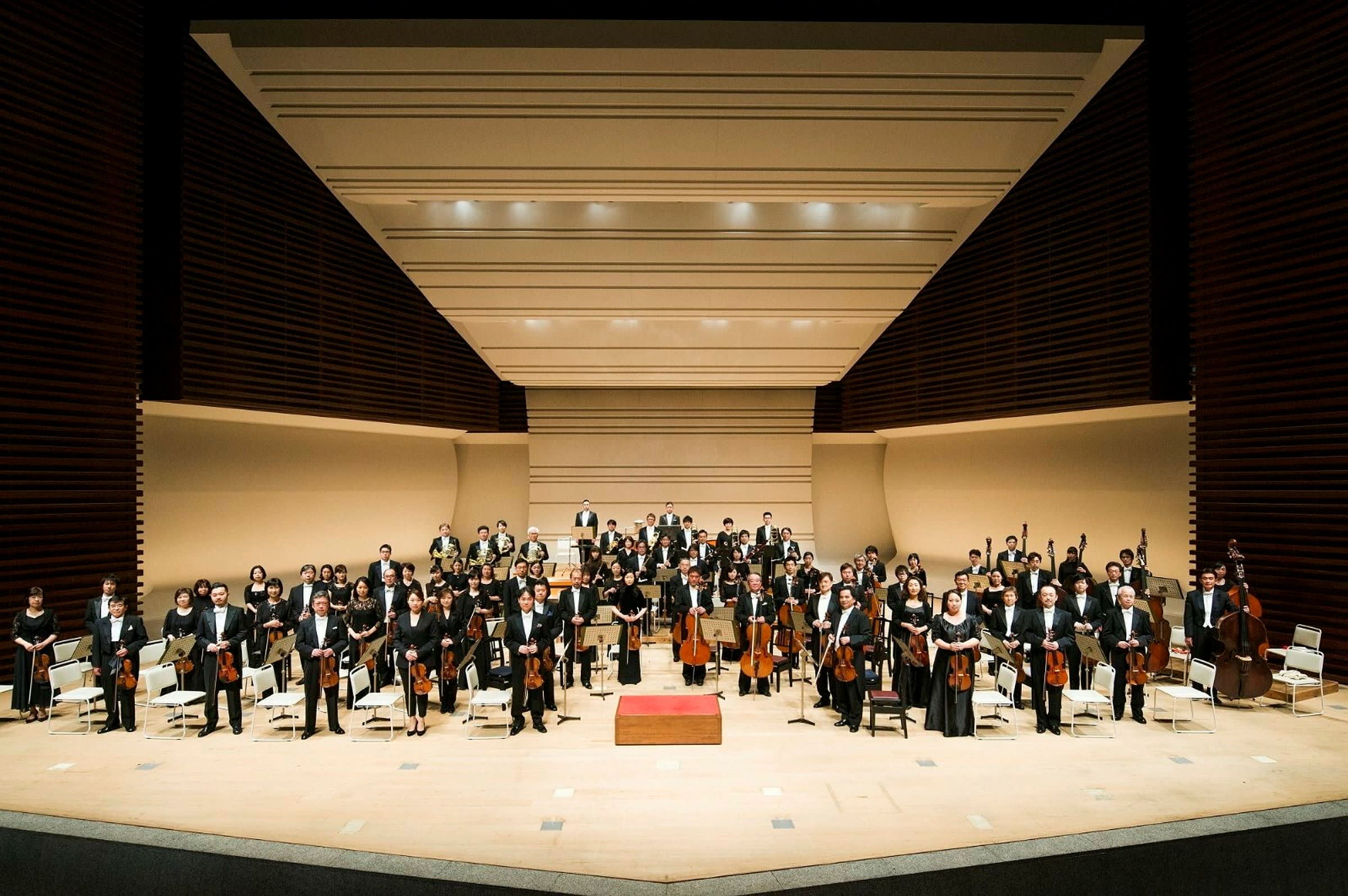 The Tokyo Metropolitan Symphony Orchestra standing on a large modernist stage