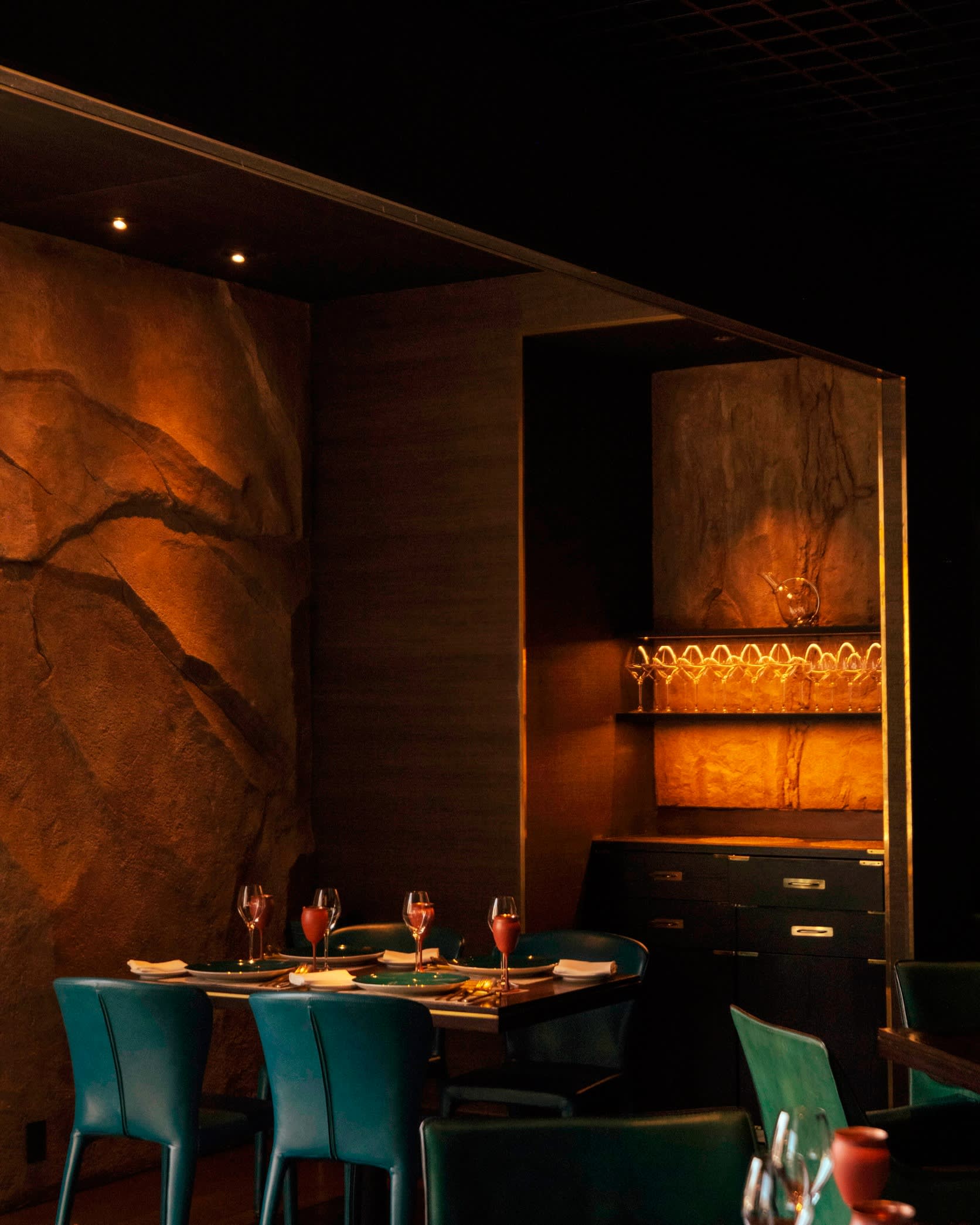 Detail of the interior of Spice Lab Tokyo, with corner table with green leather chairs by a dark-orange irregular stone wall 