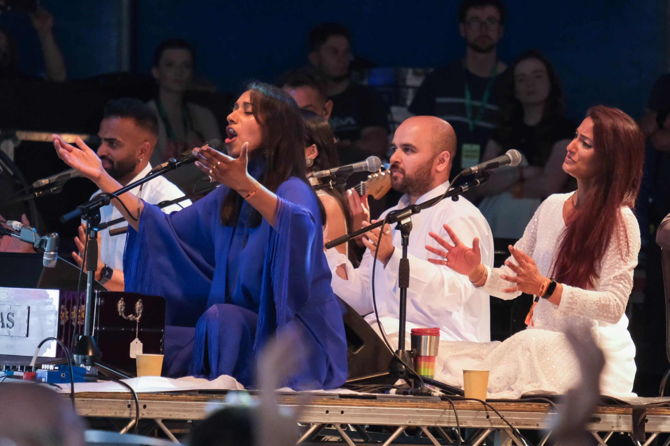 A group of male and female singers sit singing into microphones, their hands raised and open