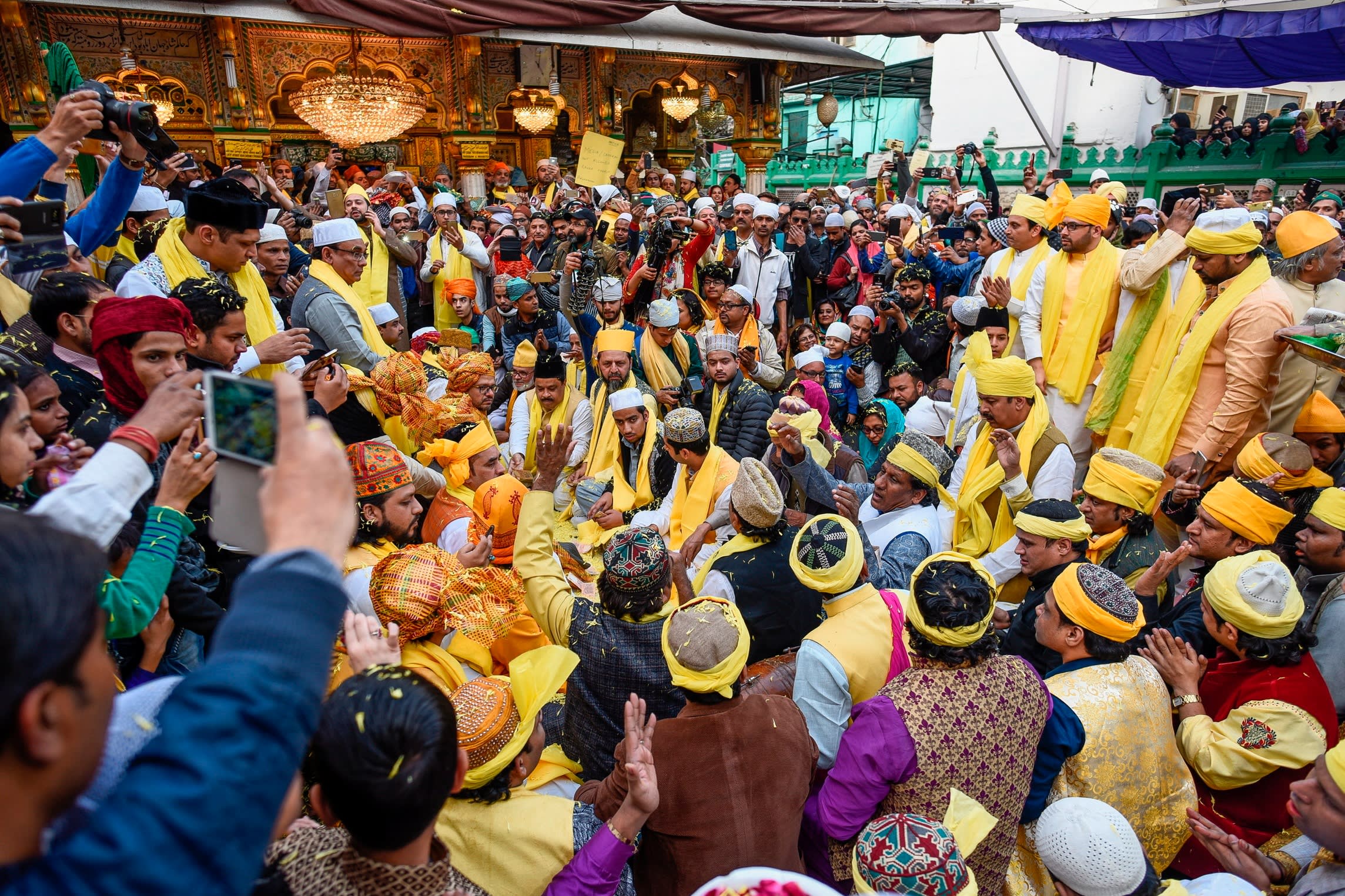 Crowds gather around a group of people seated on the ground wearing bright yellow robes, hats and turbans