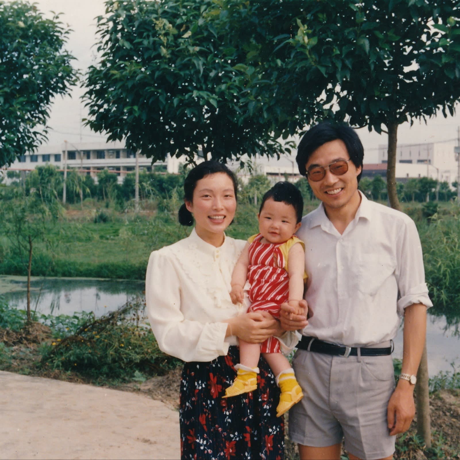 A woman in white blouse and skirt, with a man in white shirt and shorts, holding a young child wearing a red stripey top and yellow footwear