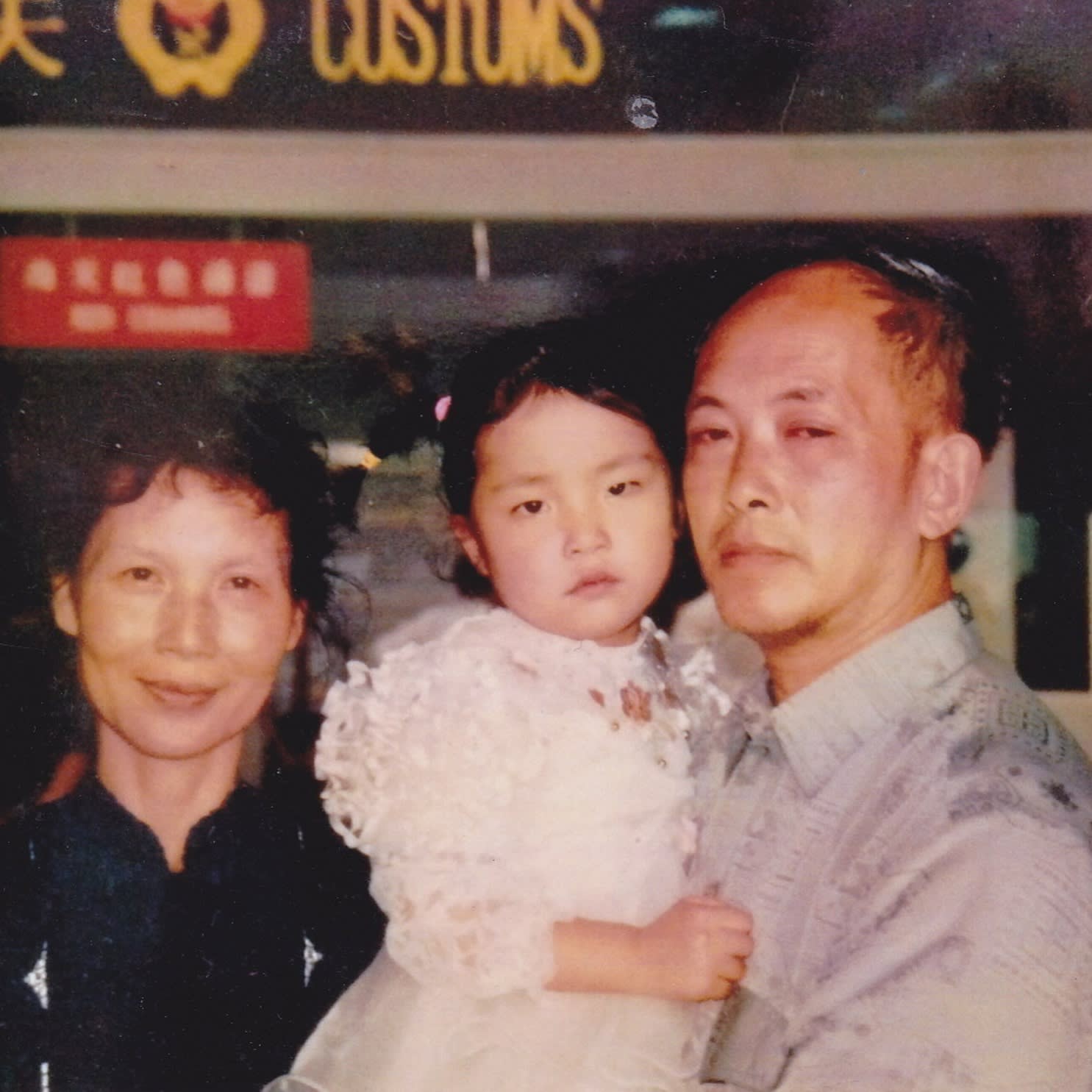A faded colour photograph of a smiling middle-aged woman in a black top and a man in a grey shirt holding a small female child in white