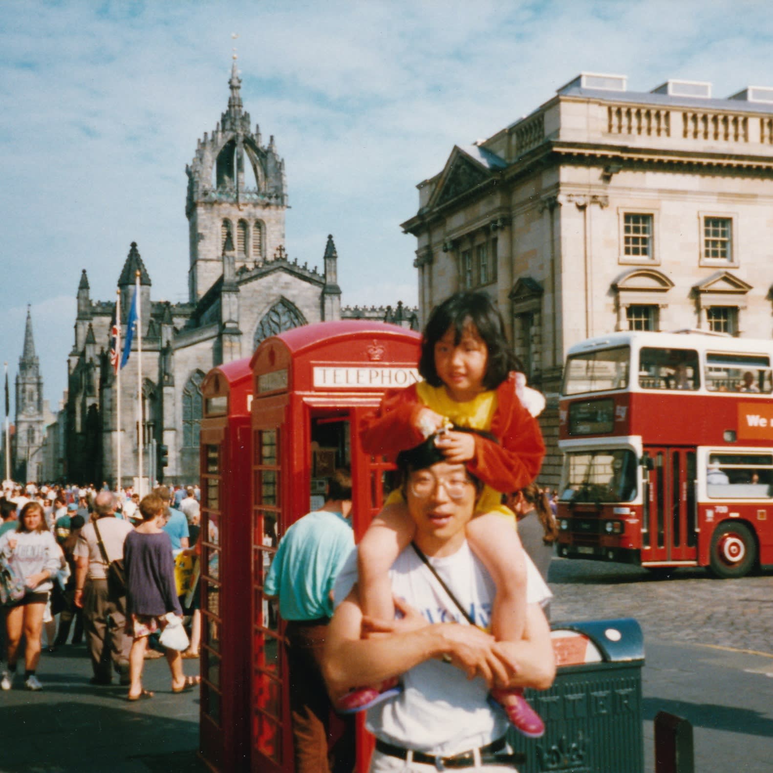 A colour photo of a four-year-old girl on the shoulders of a bespectacled man in a T-shirt standing in front of a British red telephone box near Edinburgh’s St Giles’s Cathedral