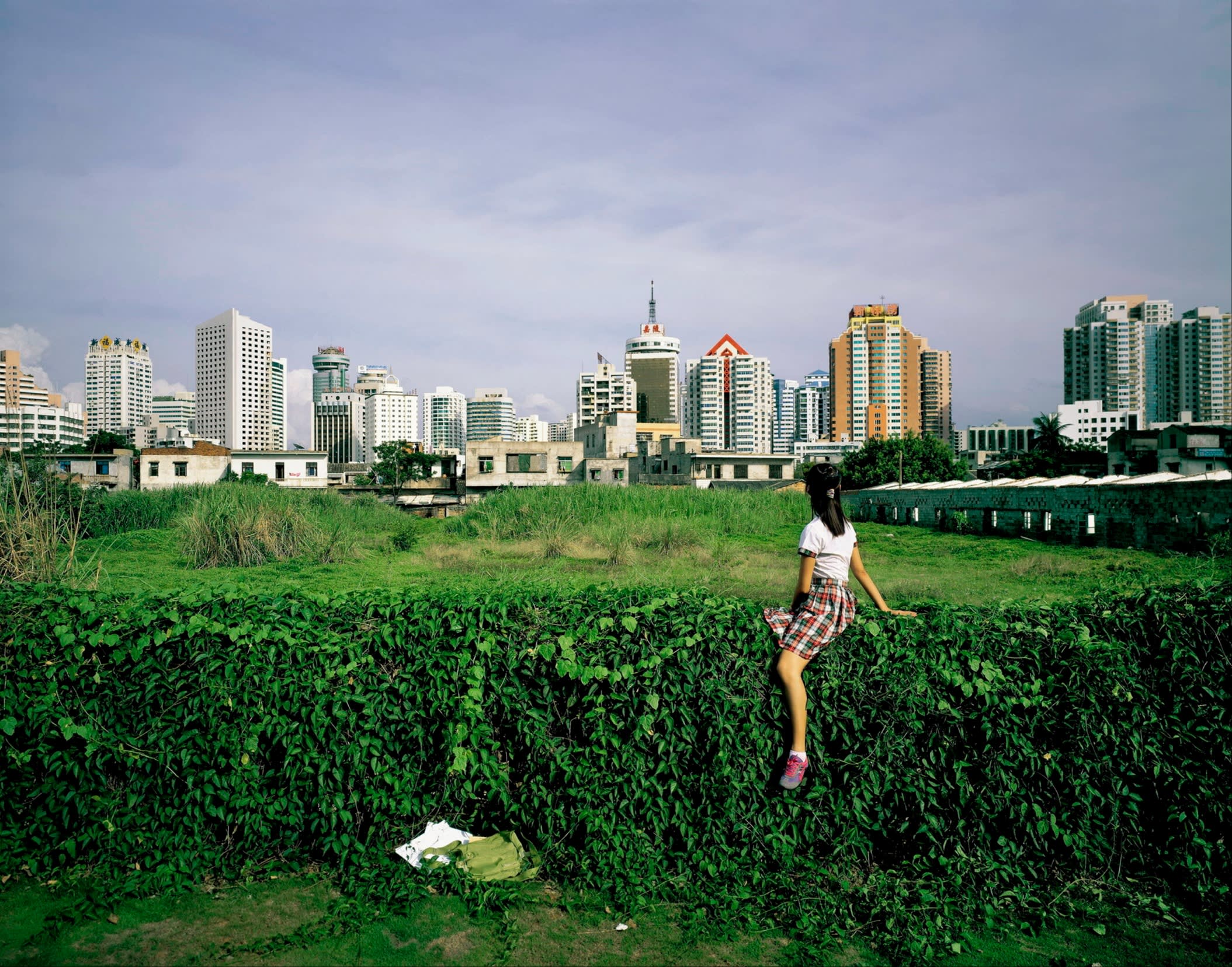 A girl or young woman with long black hair, white blouse, skirt and trainers sits on a hedge, facing across a green field at a skyline of tower blocks