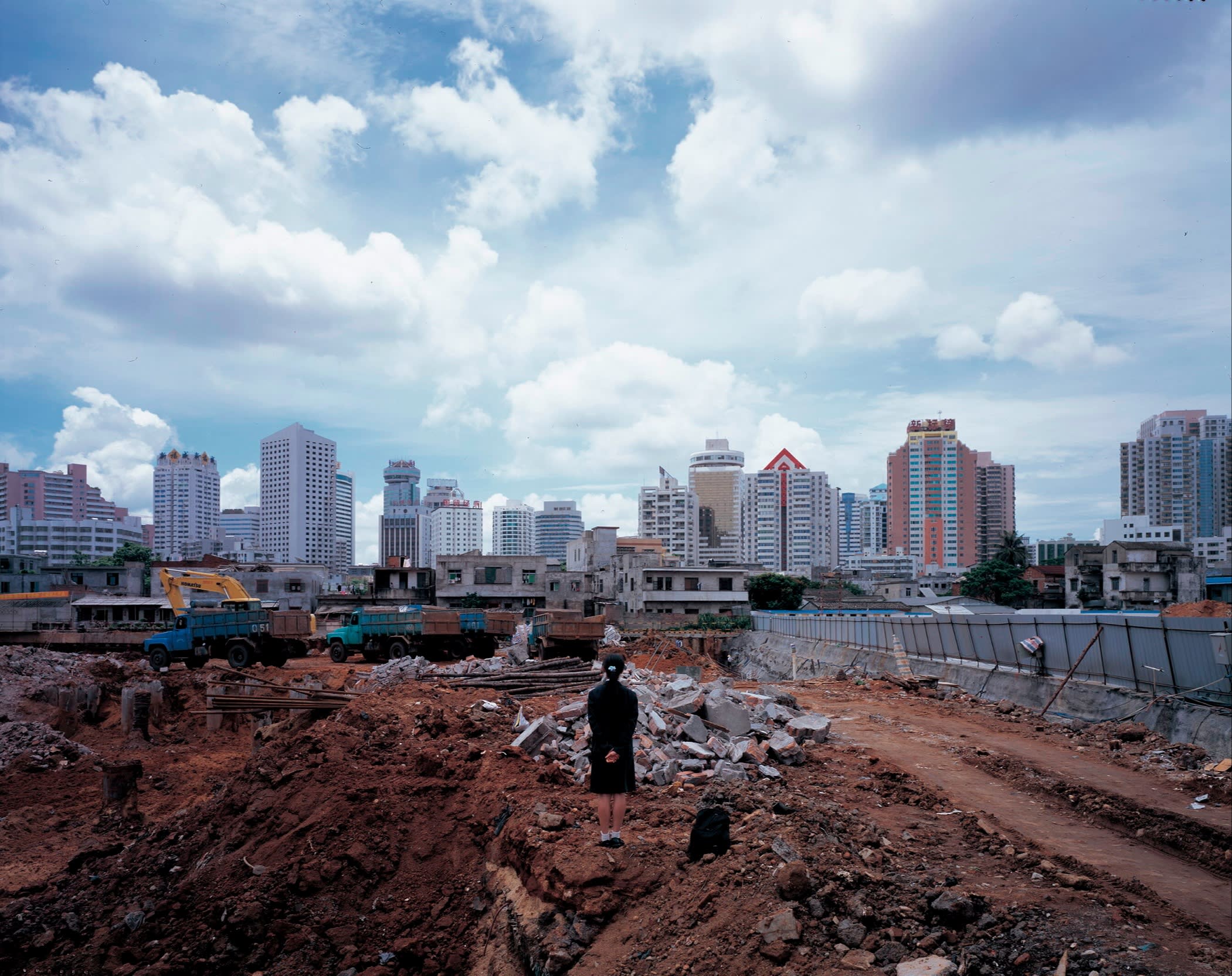 A young woman in black top and skirt stands back to the camera on a building site facing a skyline of new tower blocks
