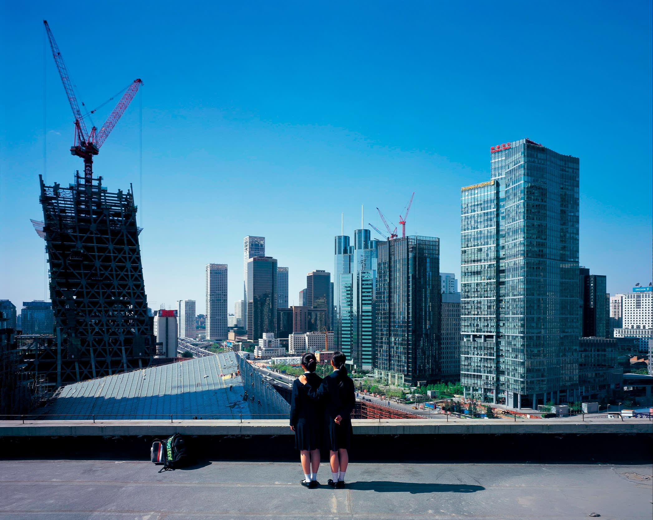 Two young women or girls in black tops and skirts stand backs to the camera facing a skyline of new tower blocks under a sunny blue sky
