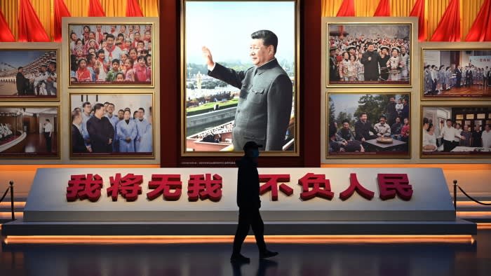 A small figure in silhouette walks past a large portrait of Xi Jinping waving. The portrait is flanked by other photographs of the Chinese president meeting the people