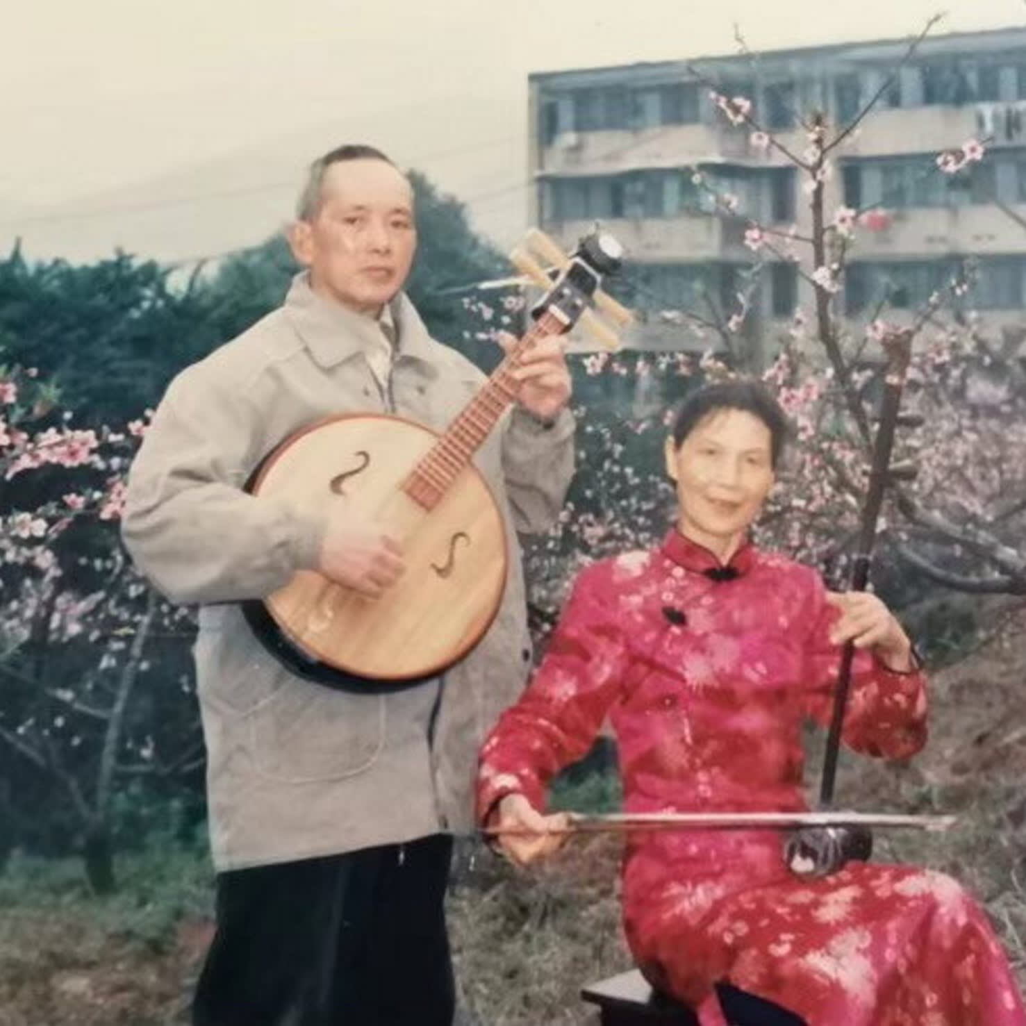 A faded colour photograph of a middle-aged man in a grey jacket and a woman in a scarlet kimono in front of a tree in blossom. The man is playing a round guitar-like or mandolin-like instrument; the woman, seated, uses a bow to play a tall, thin stringed instrument
