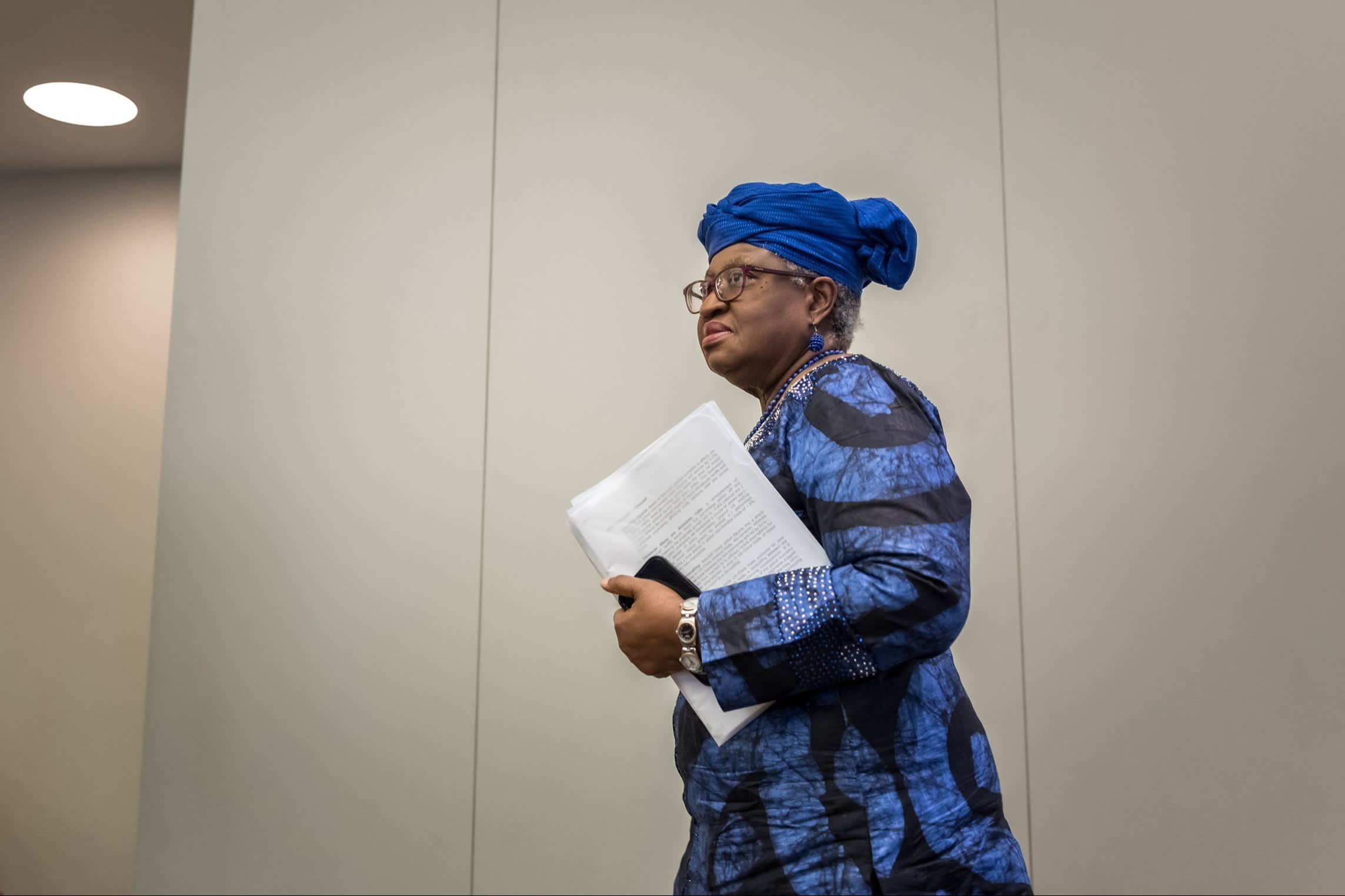 A woman wearing a blue patterned outfit and headwrap holds documents while walking indoors