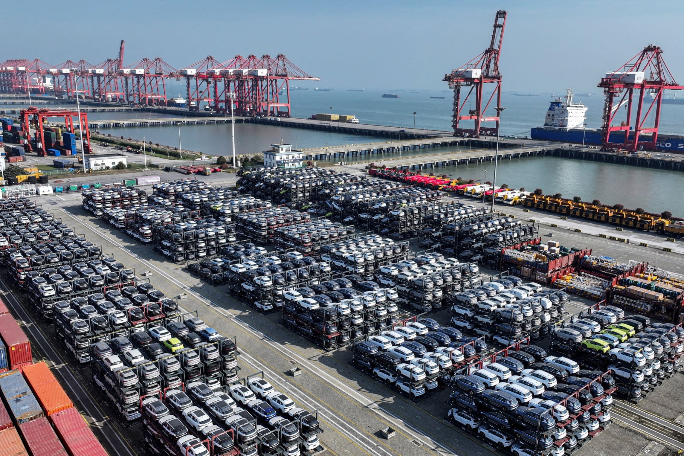 Rows of vehicles parked at a busy shipping port with large red cranes and cargo ships in the background