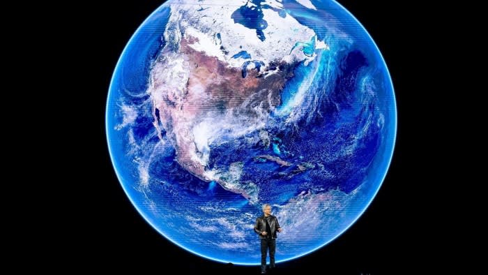 A man stands on a stage giving a talk in front of a giant screen image of a blue planet earth surrounded by the darkness of space