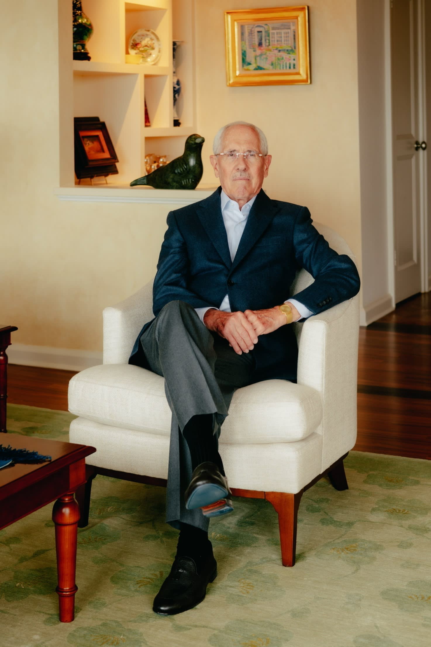 A man in a dark blue blazer and grey trousers sits on a white armchair with artworks including a sculpture of a bird behind him