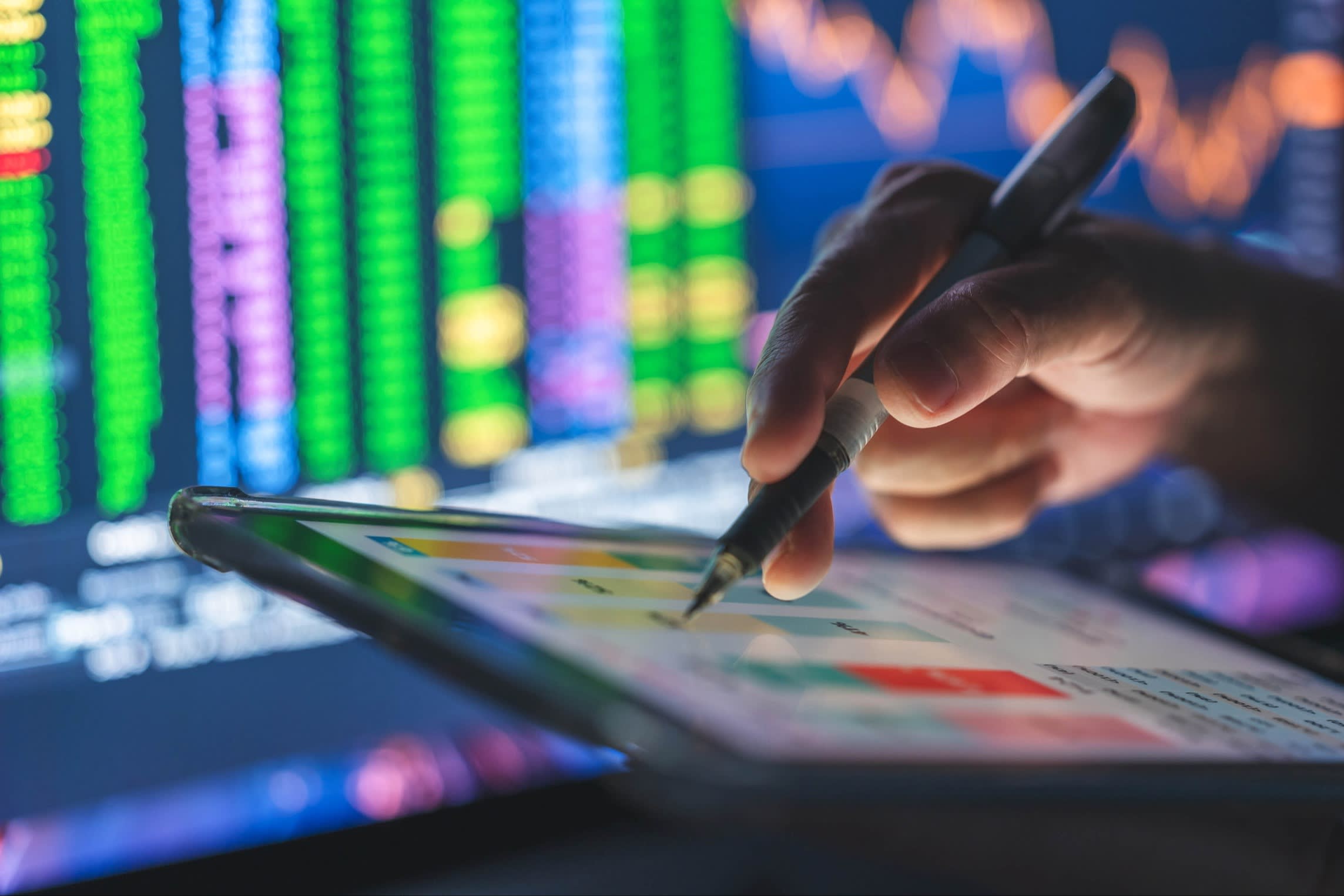 Close-up of a hand using a stylus on a tablet, with colorful stock market data on screens in the background