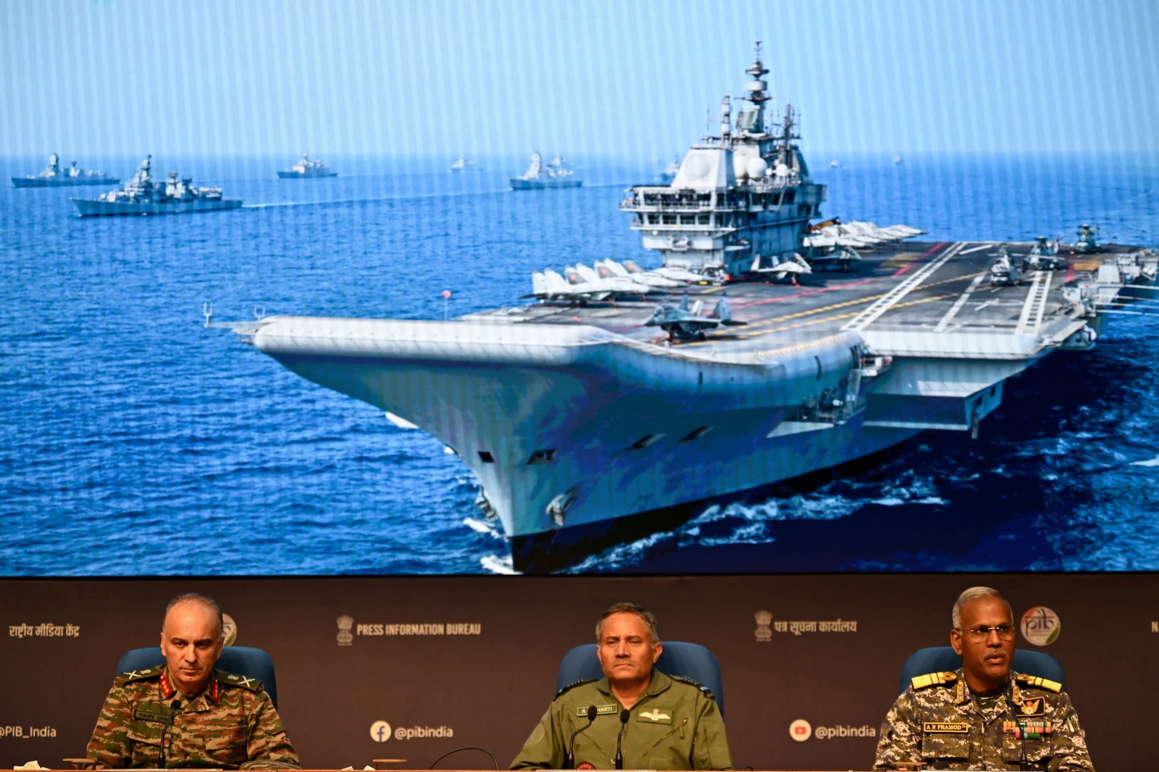Senior Indian officials sitting below a large screen showing an aircraft carrier