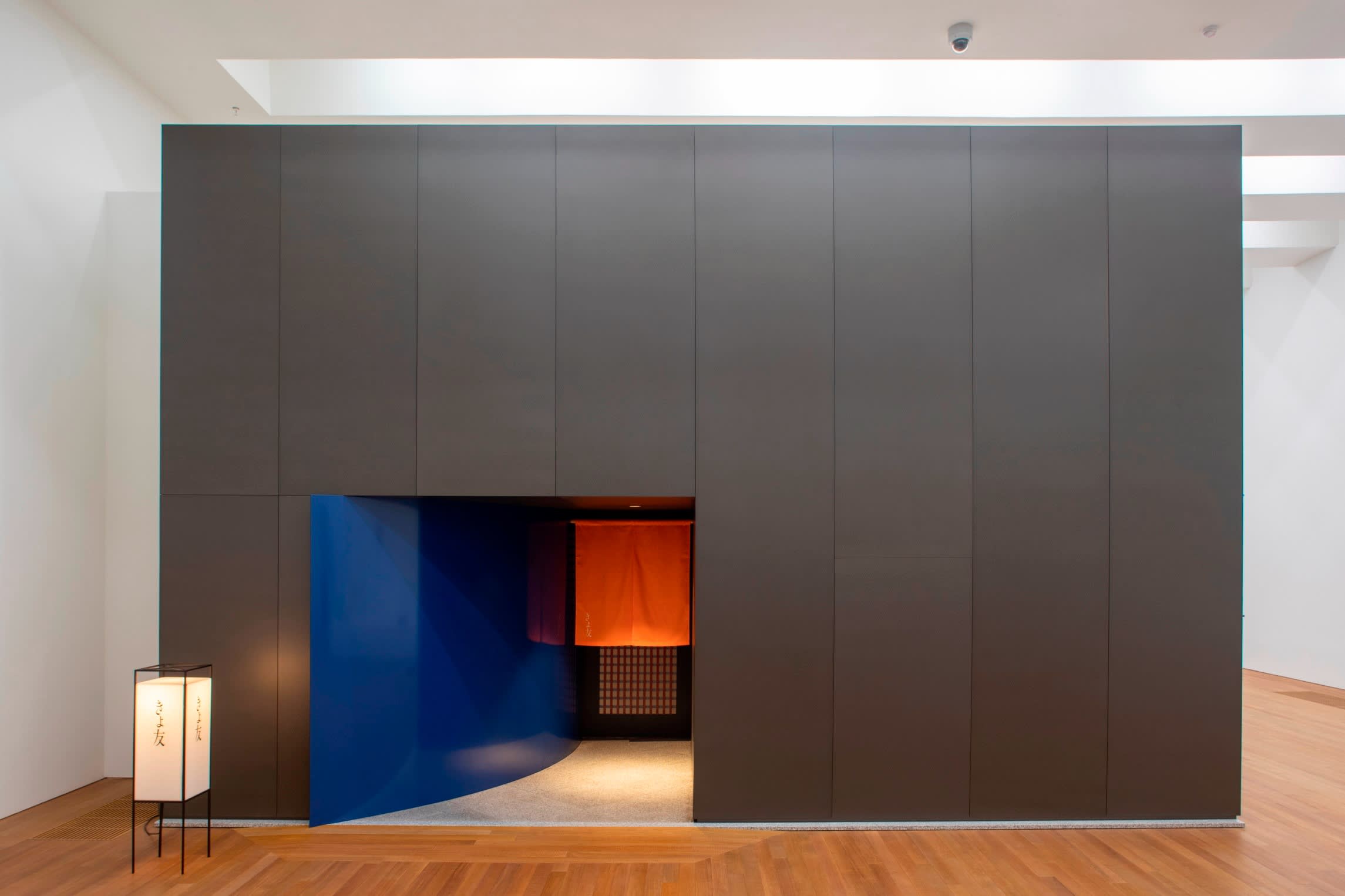 Black cube-like structure in an art gallery, with white and red paper lanterns either side of a low doorway coloured blue.