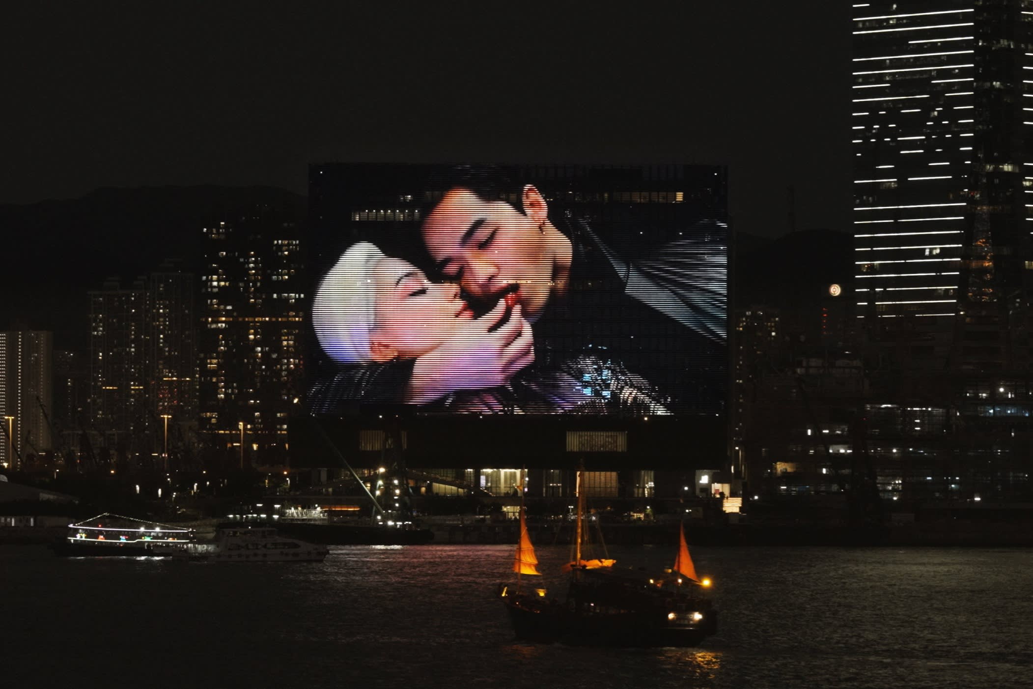 A still from what looks like an Asian movie is projected on the side of a riverside building. The image is of a handsome dark-haired young man eating a red cherry as he embraces a platinum-haired young woman