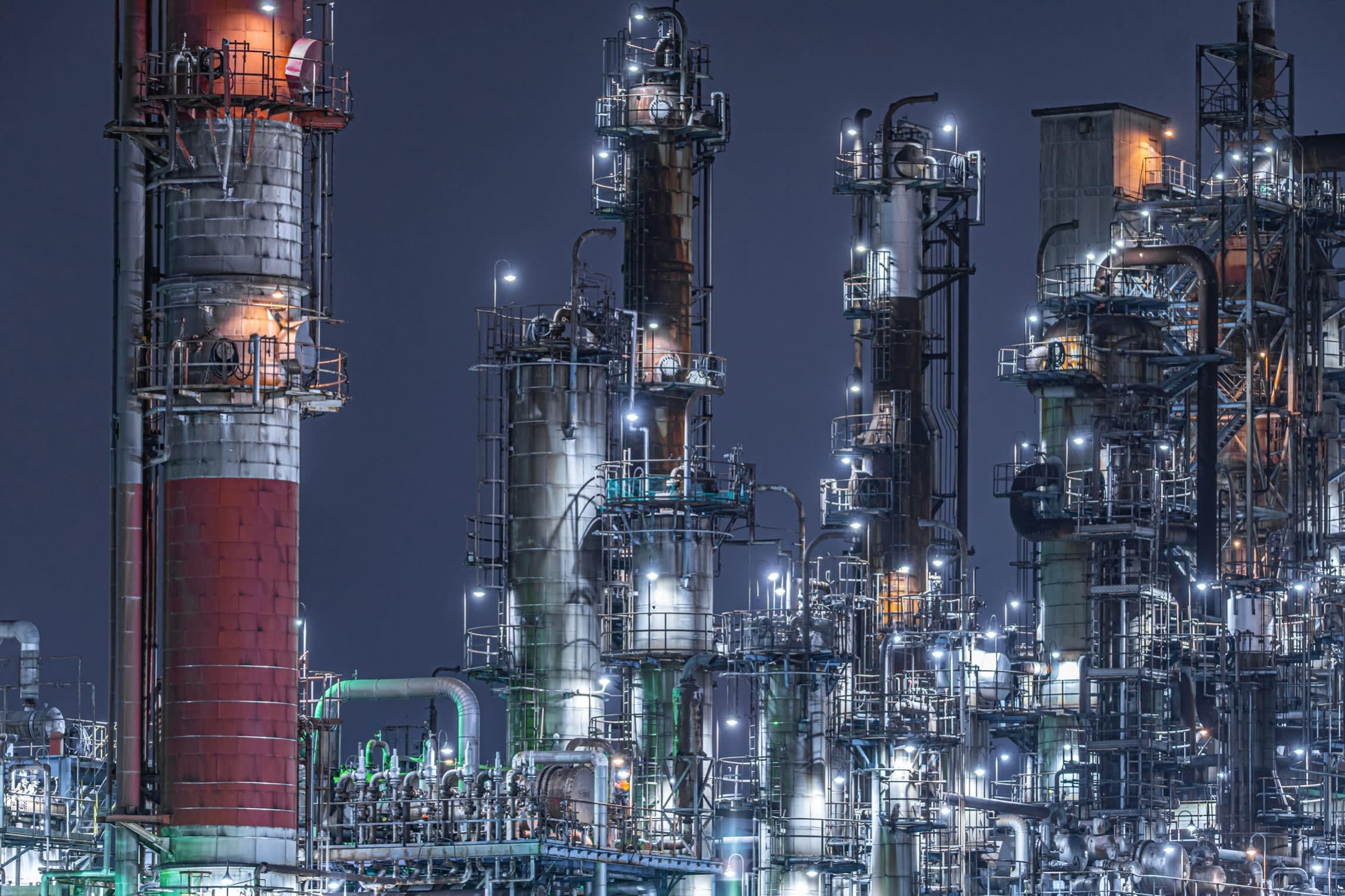 A chemical plant at night, with a large chimney and a maze of pipes and gantries floodlit against a dark sky