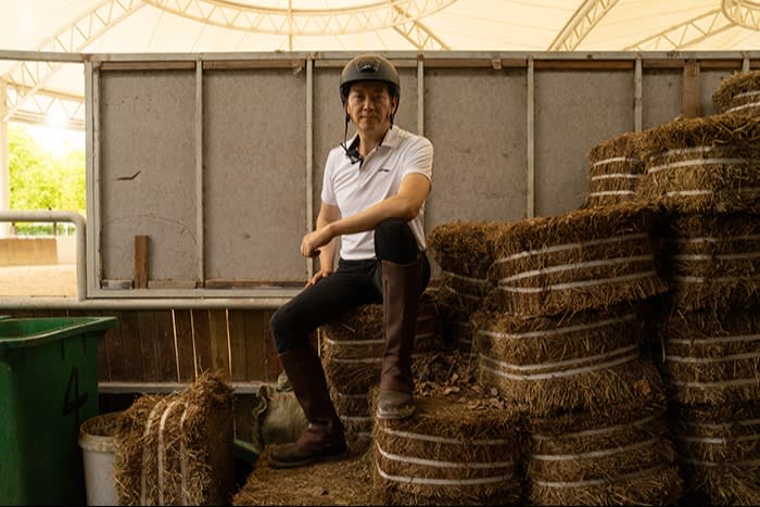 A man in equestrian gear sits comfortably atop piles of neatly stacked hay in a riding facility