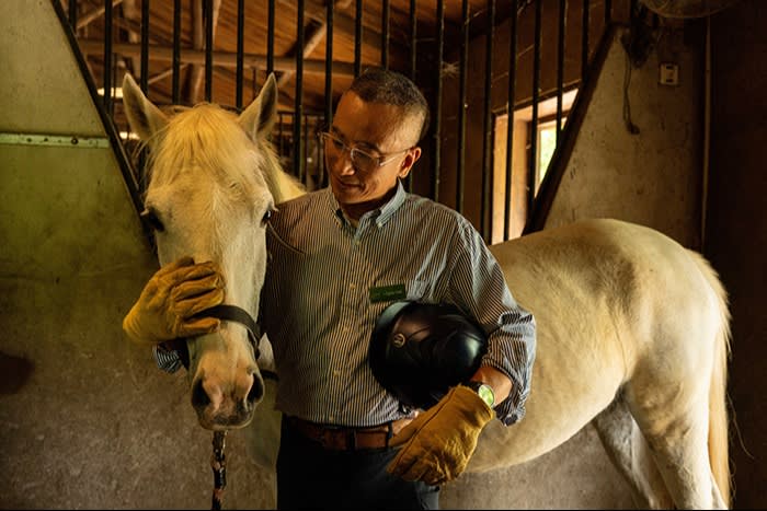 A man wearing gloves gently pets the face of a white horse inside a softly lit stable