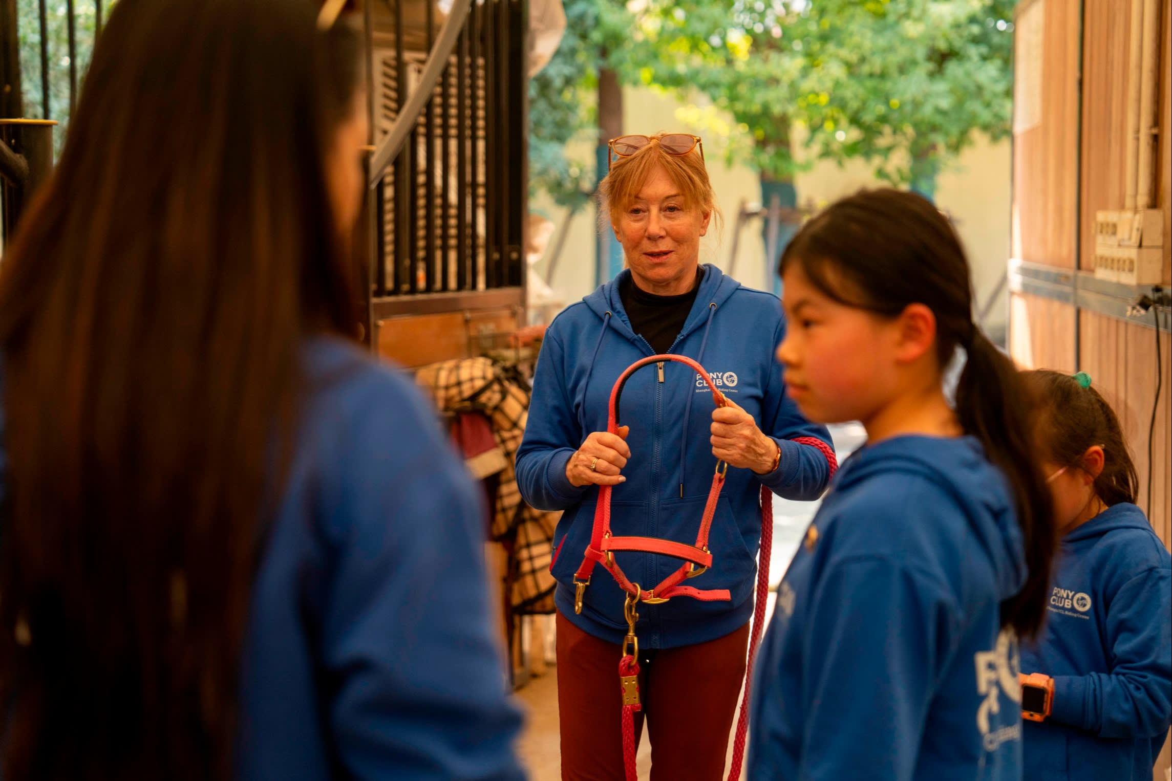 An instructor holding a pink horse halter is speaking to a group of young equestrian students wearing blue hoodies