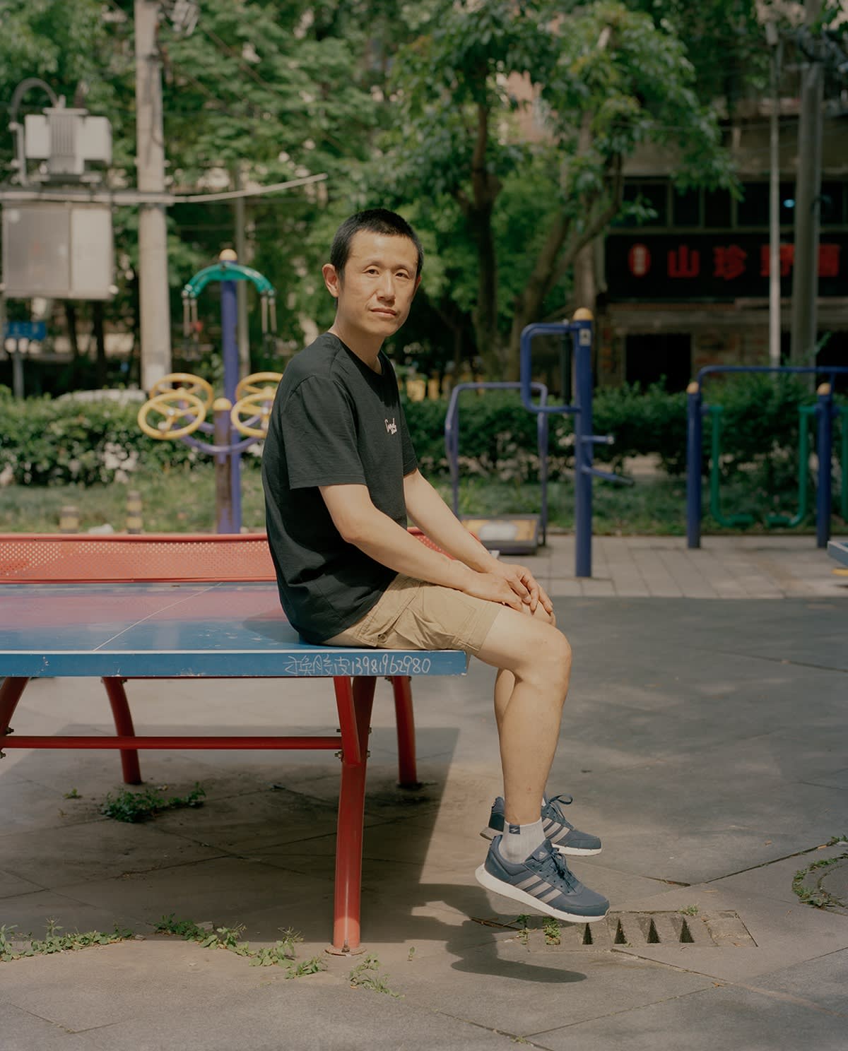 “A man sits on a ping-pong table in an outdoor park, looking at the camera