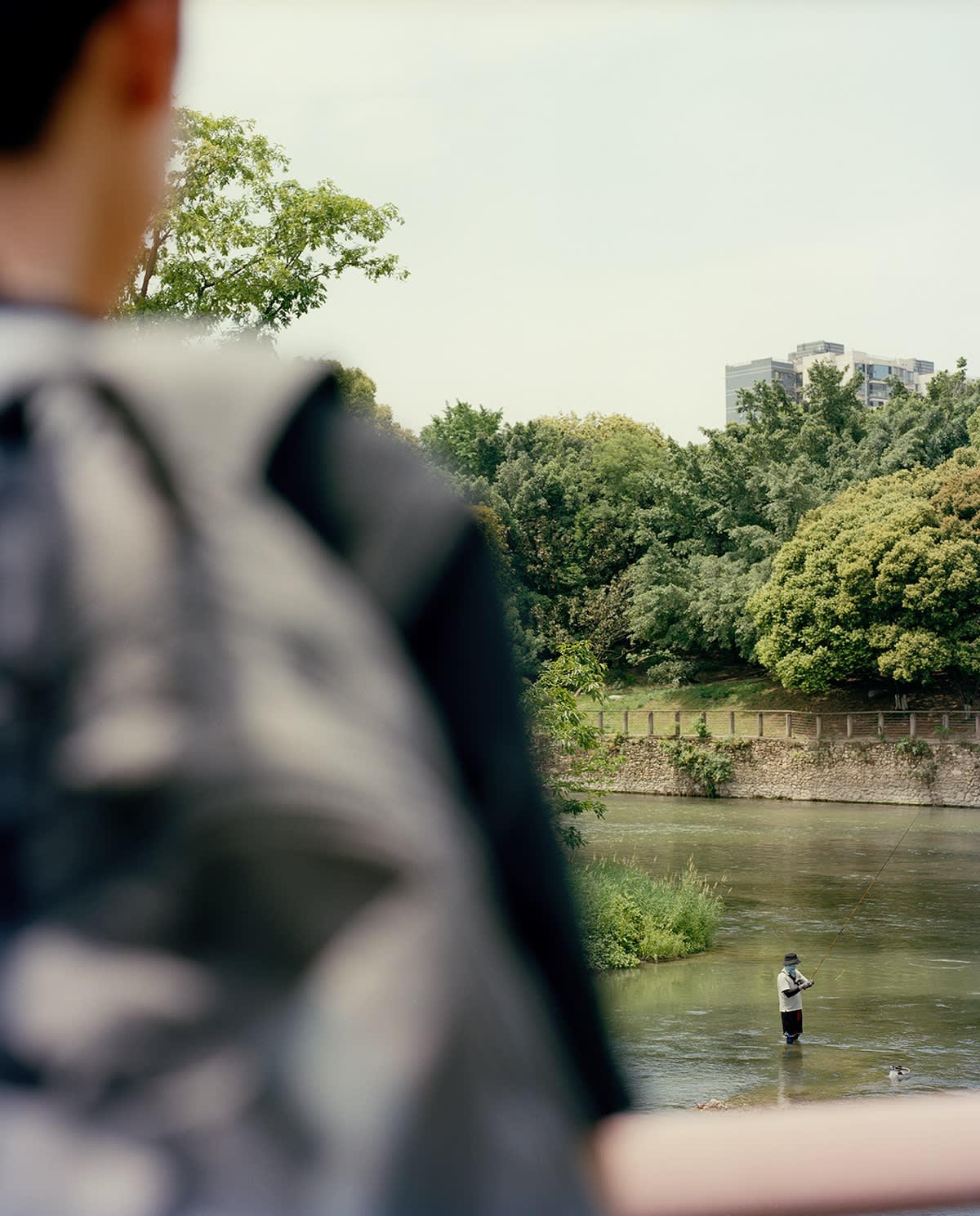 A person with a backpack observes someone fishing in a river