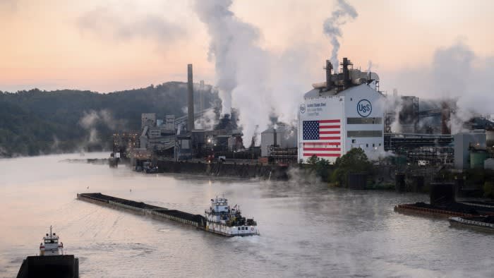 A tugboat pushes a barge along a river near the US Steel Corp facility in Clairton. The industrial site emits smoke, and an American flag is displayed on a building.