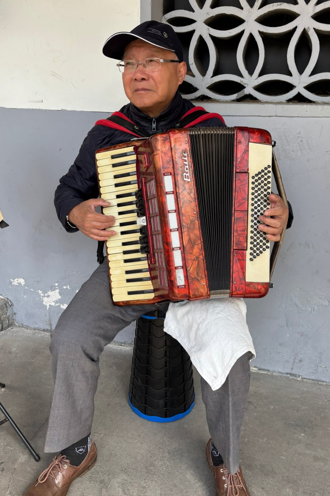 An elderly man sits on a stool playing an accordion. He is wearing a black cap, glasses, and a dark jacket
