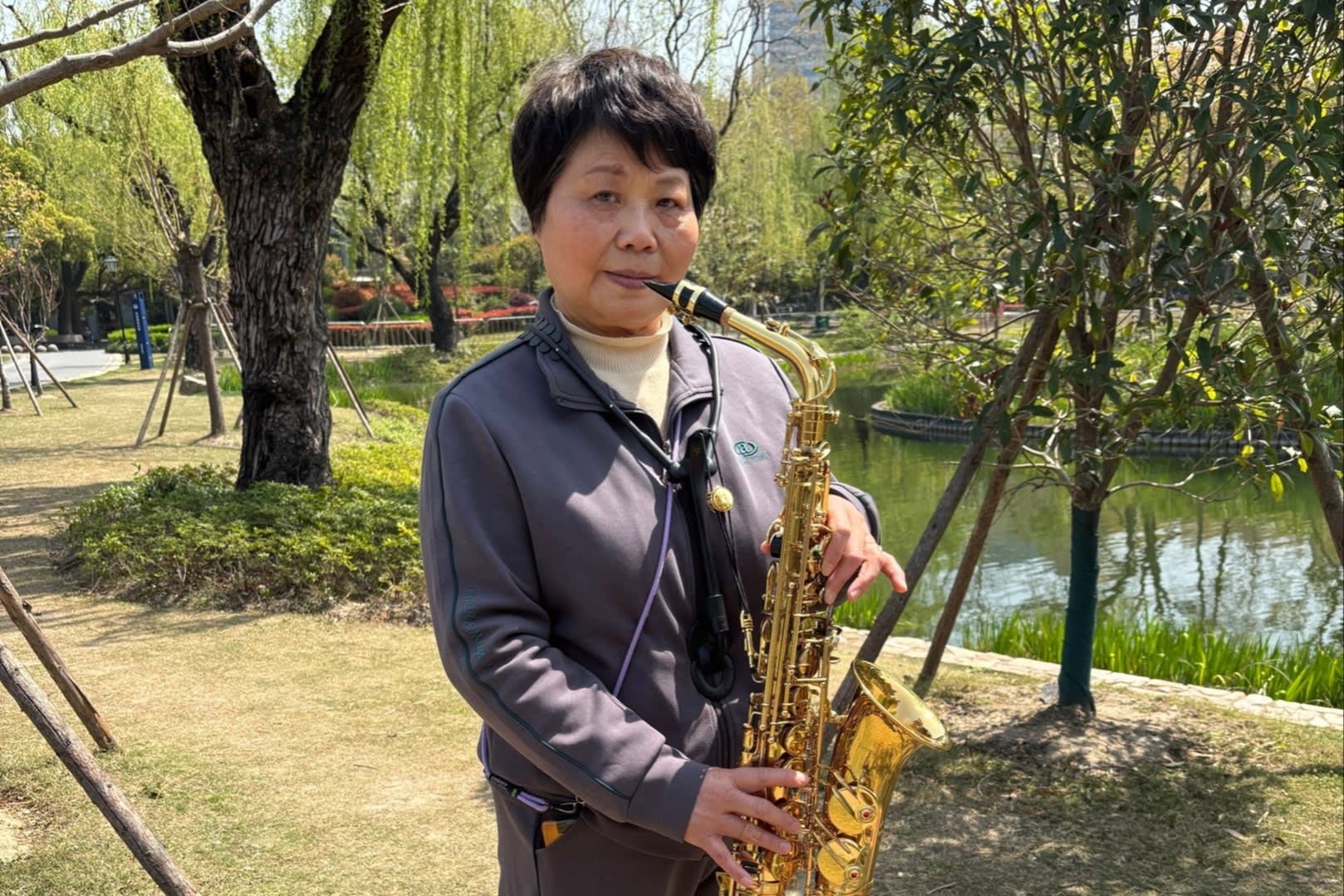Li Duping stands in a park holding a saxophone. She is dressed in a dark tracksuit, with trees and a pond in the background.