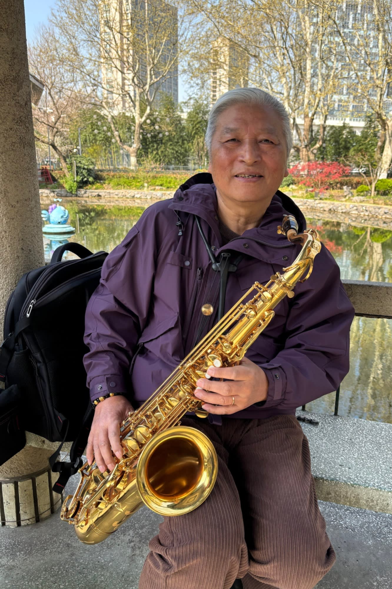 Ma Zhiqiang holds a saxophone. He is wearing a purple jacket and is surrounded by a serene park setting with trees and buildings in the background