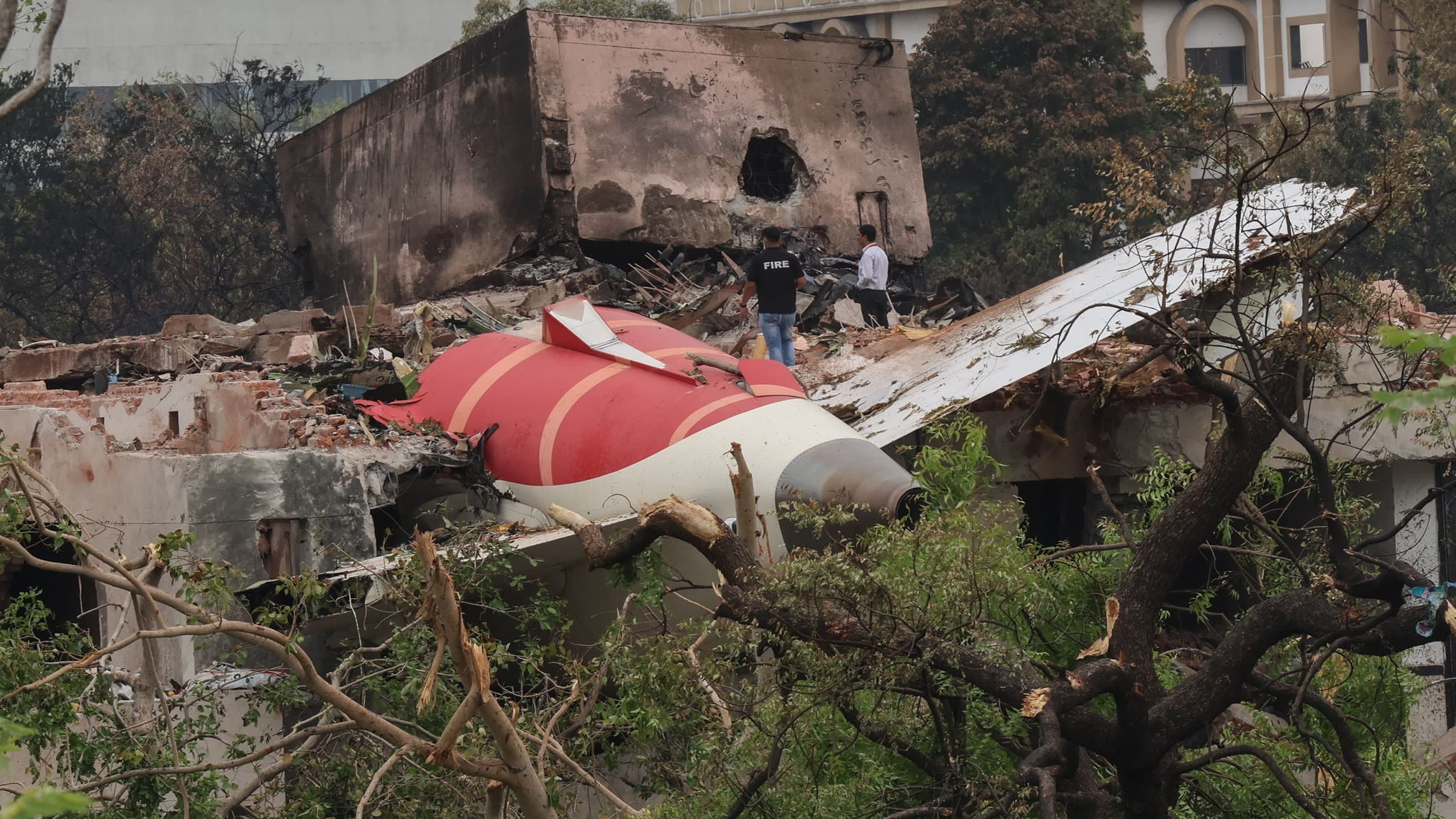 Debris from the Air India plane is scattered near a damaged building. Two officials are inspecting the crash site amid the wreckage and fallen trees