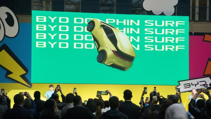A crowd gathers at the launch event for the BYD Dolphin Surf electric vehicle. The audience is taking photos and videos of a large screen displaying an image of the yellow car
