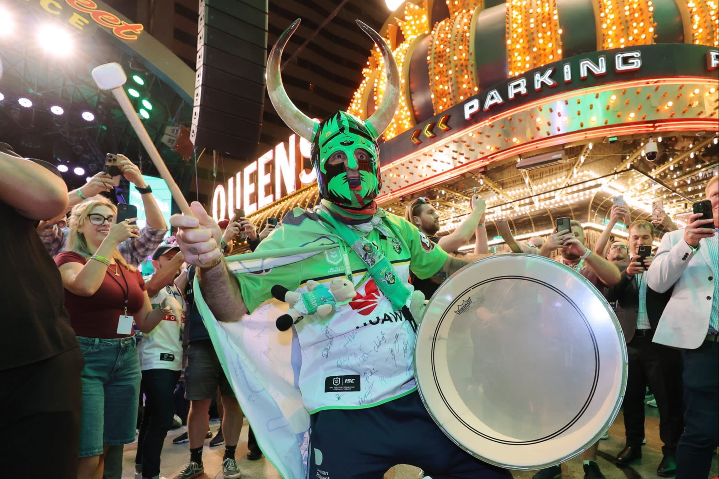 A man wearing a green mask and horned helmet bangs a drum during a parade past a brightly lit Las Vegas venue