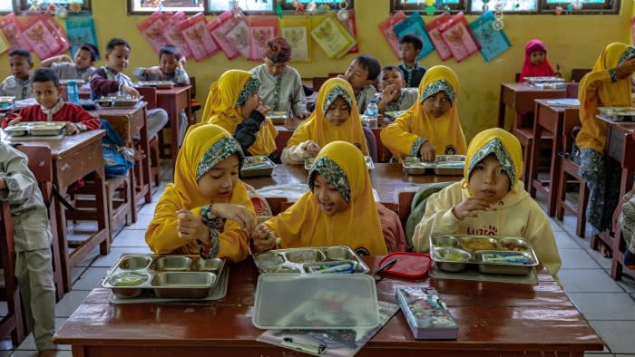 A classroom is filled with children, some wearing yellow hijabs, as they eat from metal trays.