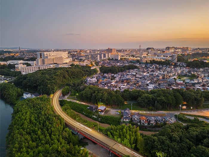 Aerial view of a city at sunset with a curved road cutting through green forest and dense residential neighbourhoods, with urban buildings in the distance
