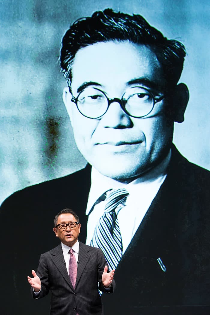 A speaker in a dark suit gestures on stage in front of a large monochrome image of an older man in glasses
